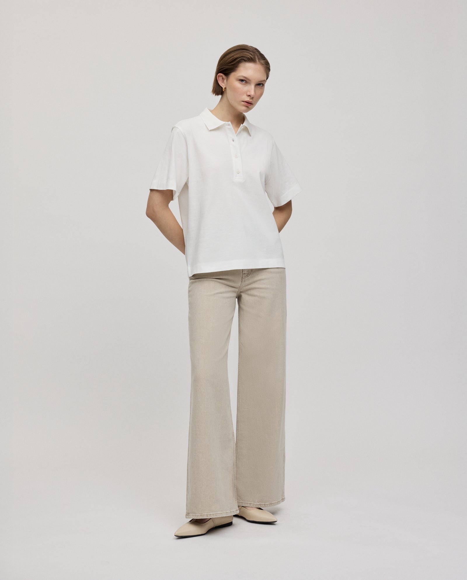A woman stands with hands in pockets, wearing the IVY OAK FABIOLA Top in white—an organic cotton short-sleeve polo shirt—paired with beige wide-leg pants.