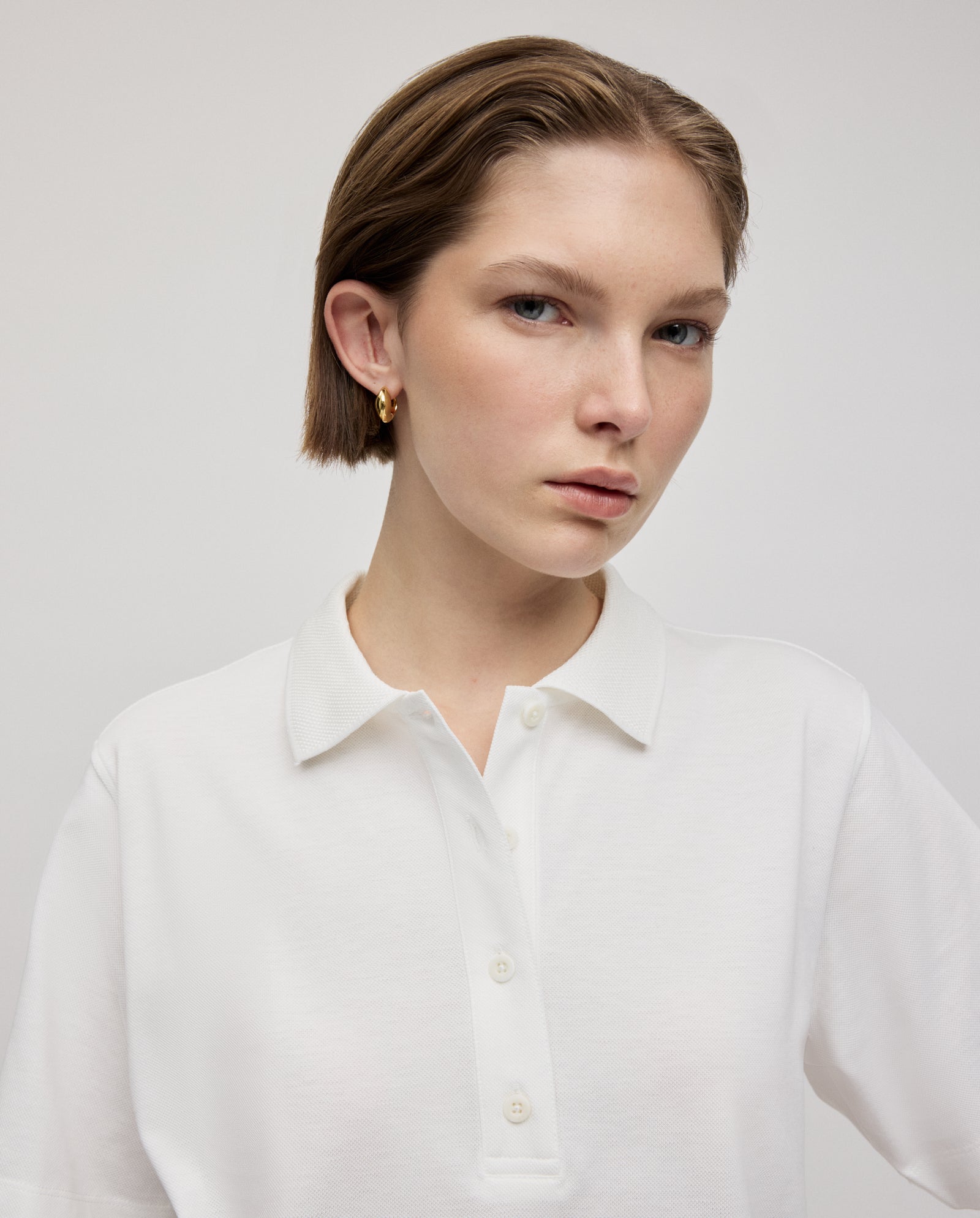 Woman with short brown hair wears the IVY OAK FABIOLA Top, a white short-sleeve polo shirt made from organic cotton, paired with gold hoop earrings against a plain background.