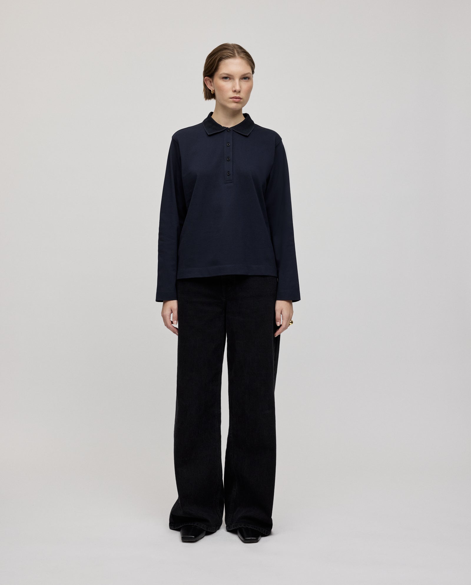 A person stands facing forward, wearing the IVY OAK FABIOLA ANN black long-sleeve top crafted from comfortable organic cotton and paired with wide-leg black pants.