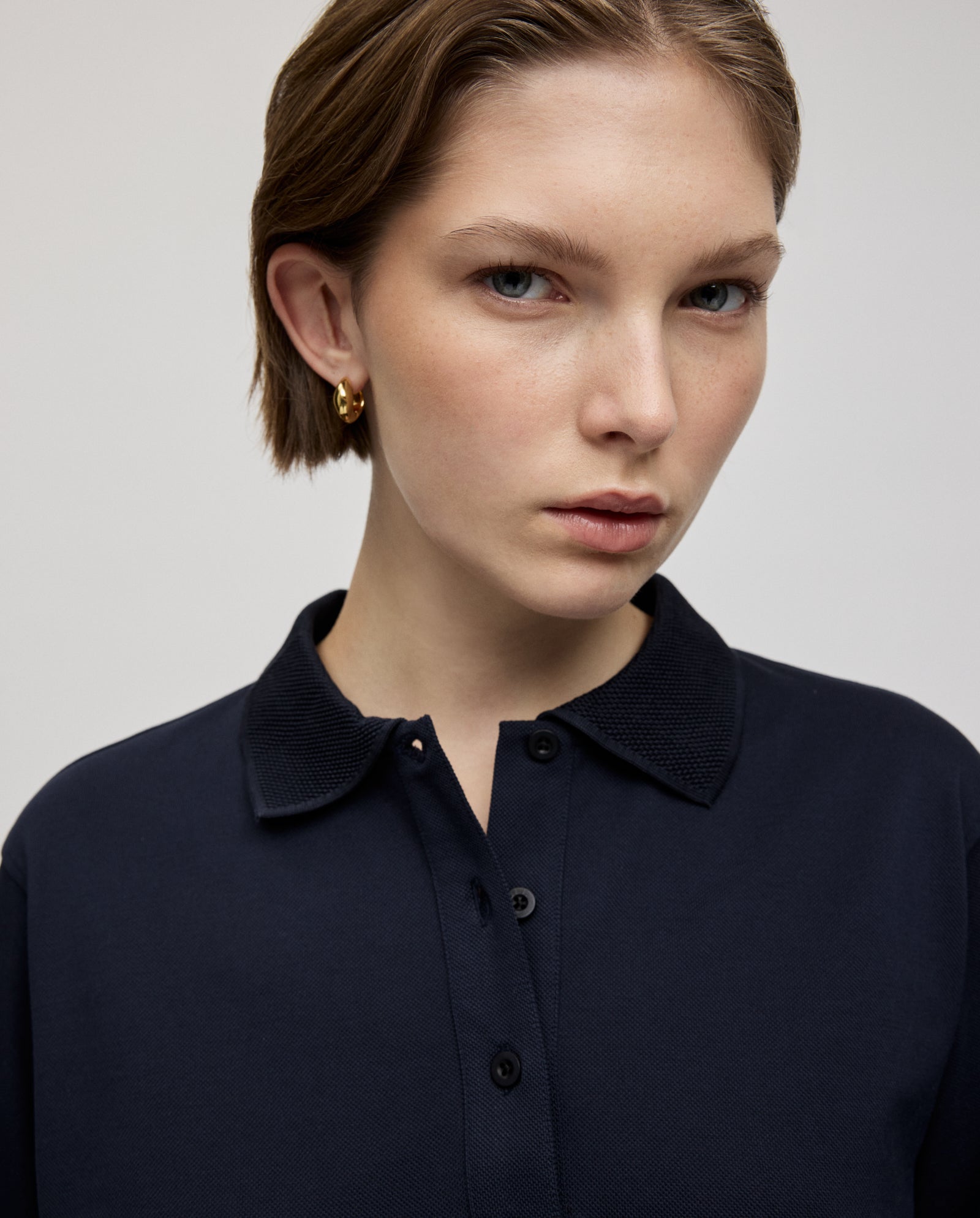 A woman with short brown hair wears the IVY OAK FABIOLA ANN Top in organic cotton and gold hoop earrings as she looks at the camera.