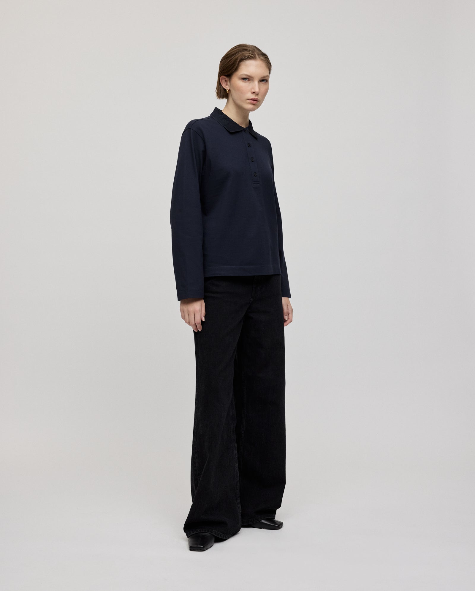 A woman wearing the IVY OAK FABIOLA ANN Top in navy, featuring long sleeves and crafted from organic cotton, paired with black wide-leg pants, stands against a plain light background.