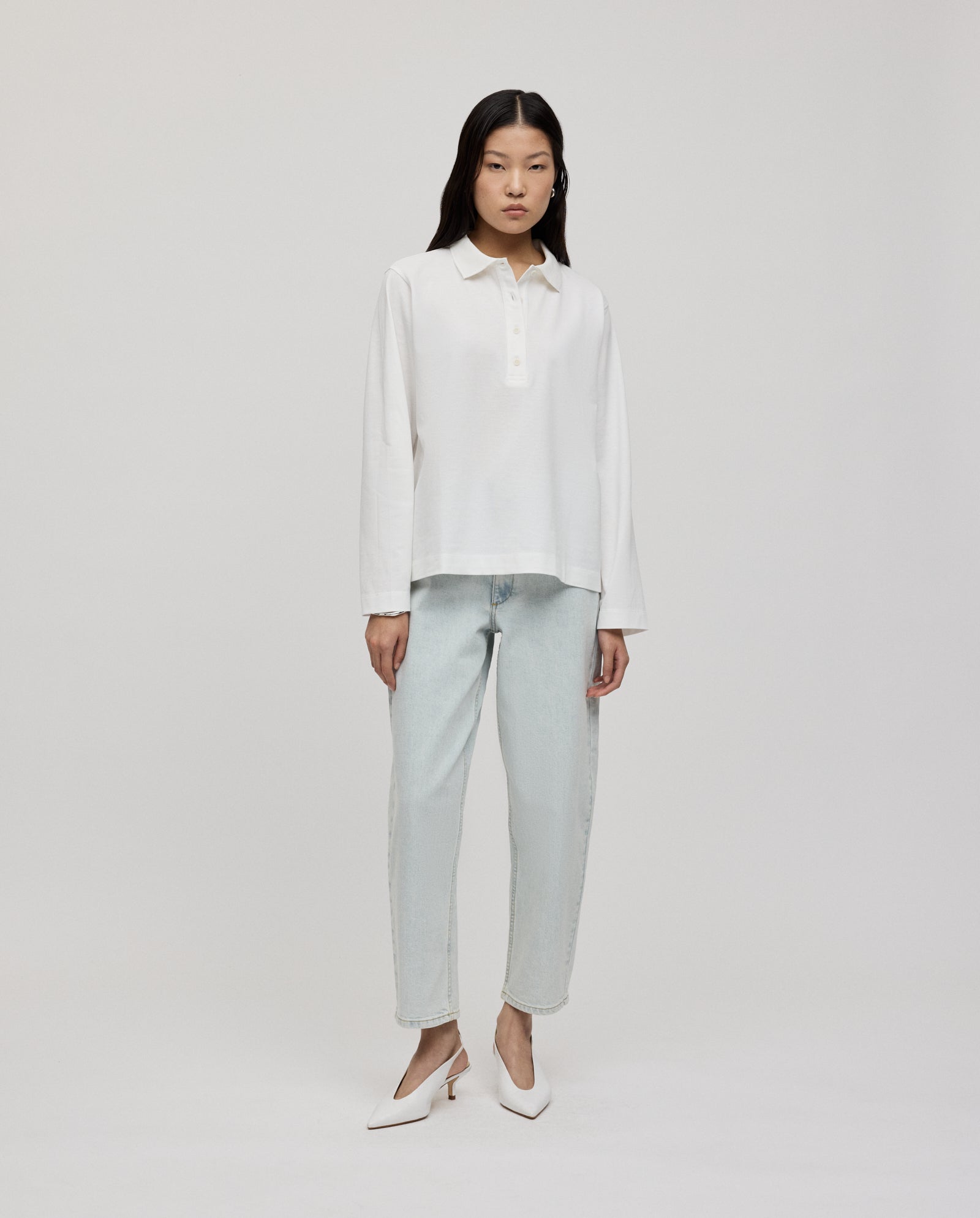 Woman standing in the IVY OAK FABIOLA ANN organic cotton long-sleeve polo shirt, light-wash jeans, and white heels against a plain background.