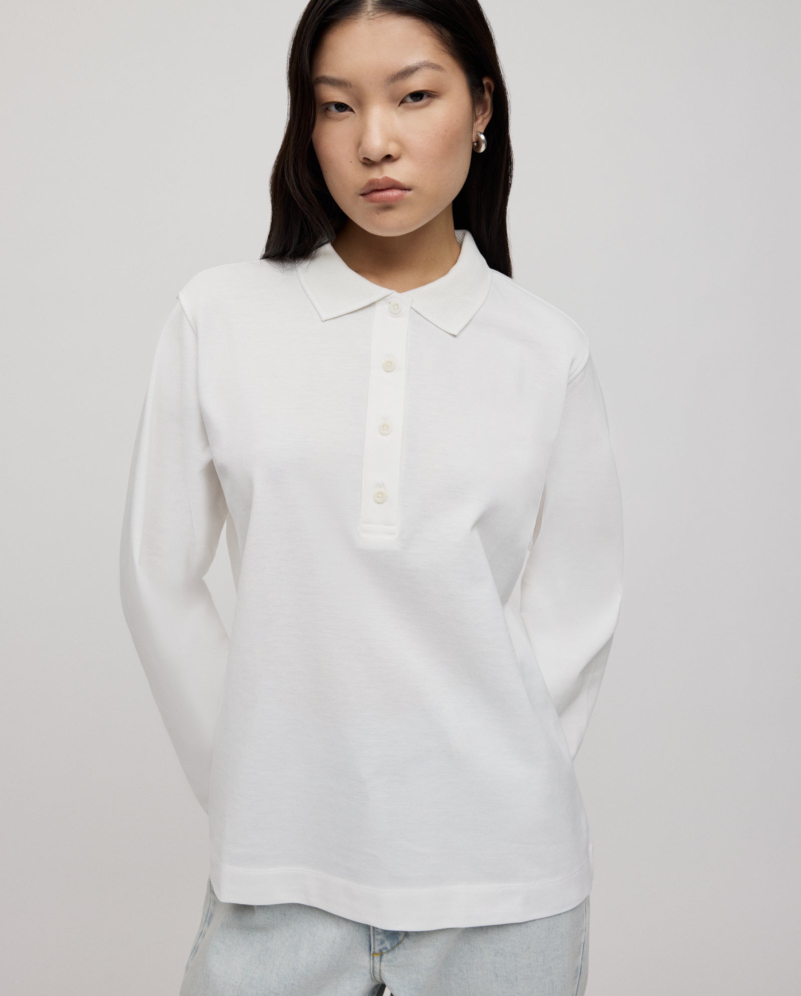 A woman with long dark hair wears the IVY OAK FABIOLA ANN Top, an organic cotton white polo shirt, paired with light jeans, standing against a plain background.