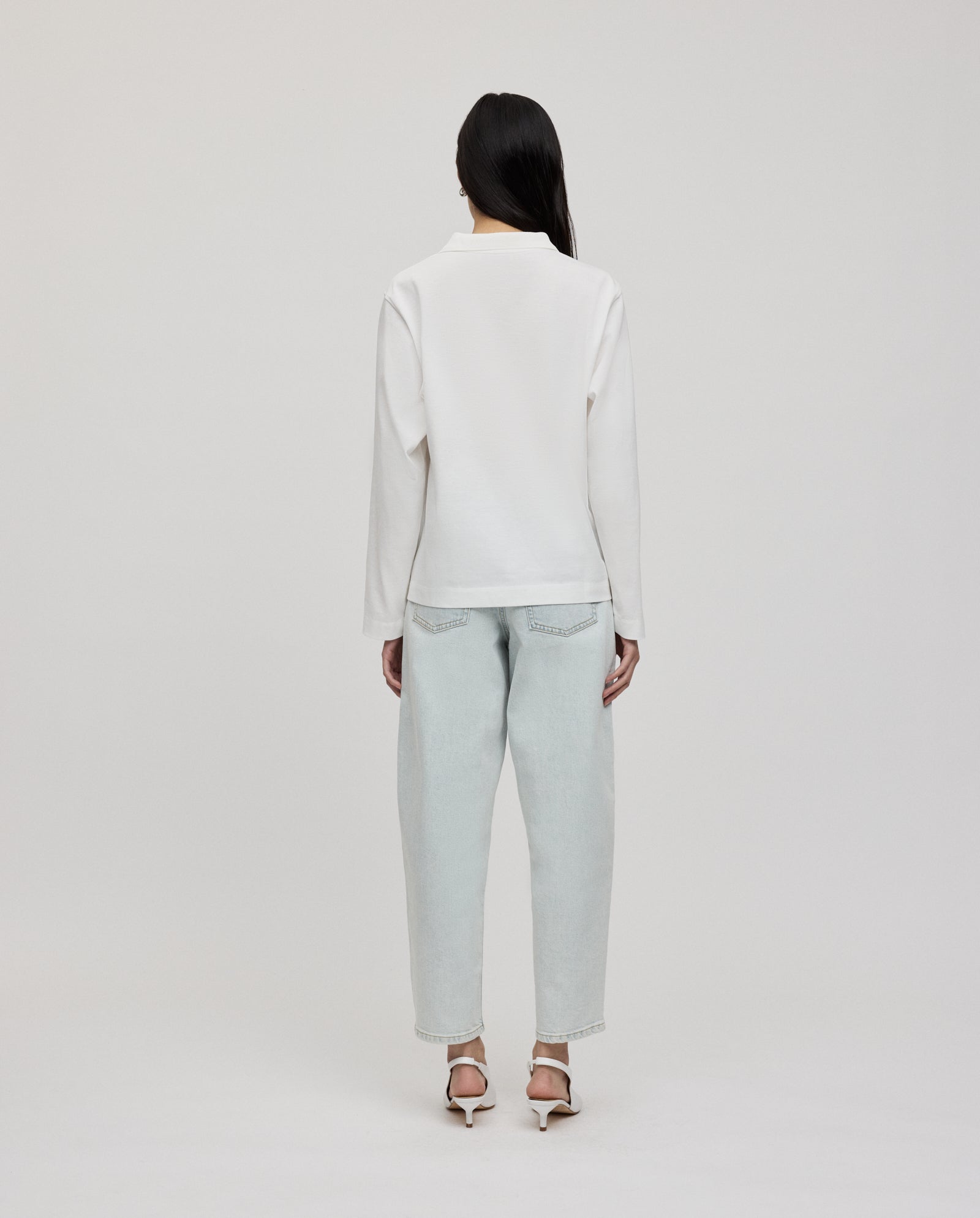 A person stands facing away against a plain background, wearing the IVY OAK FABIOLA ANN Top in white organic cotton, paired with light jeans and white heels.