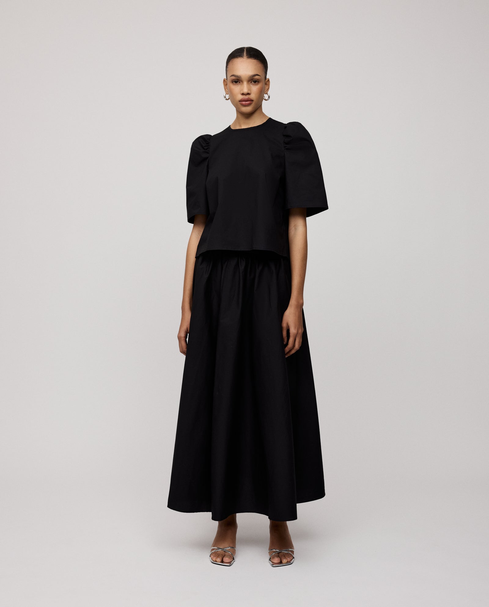 A woman wears the IVY OAK ELISABETH ANN blouse, featuring a cropped cotton fit and voluminous sleeves, paired with a long black skirt against a plain background.