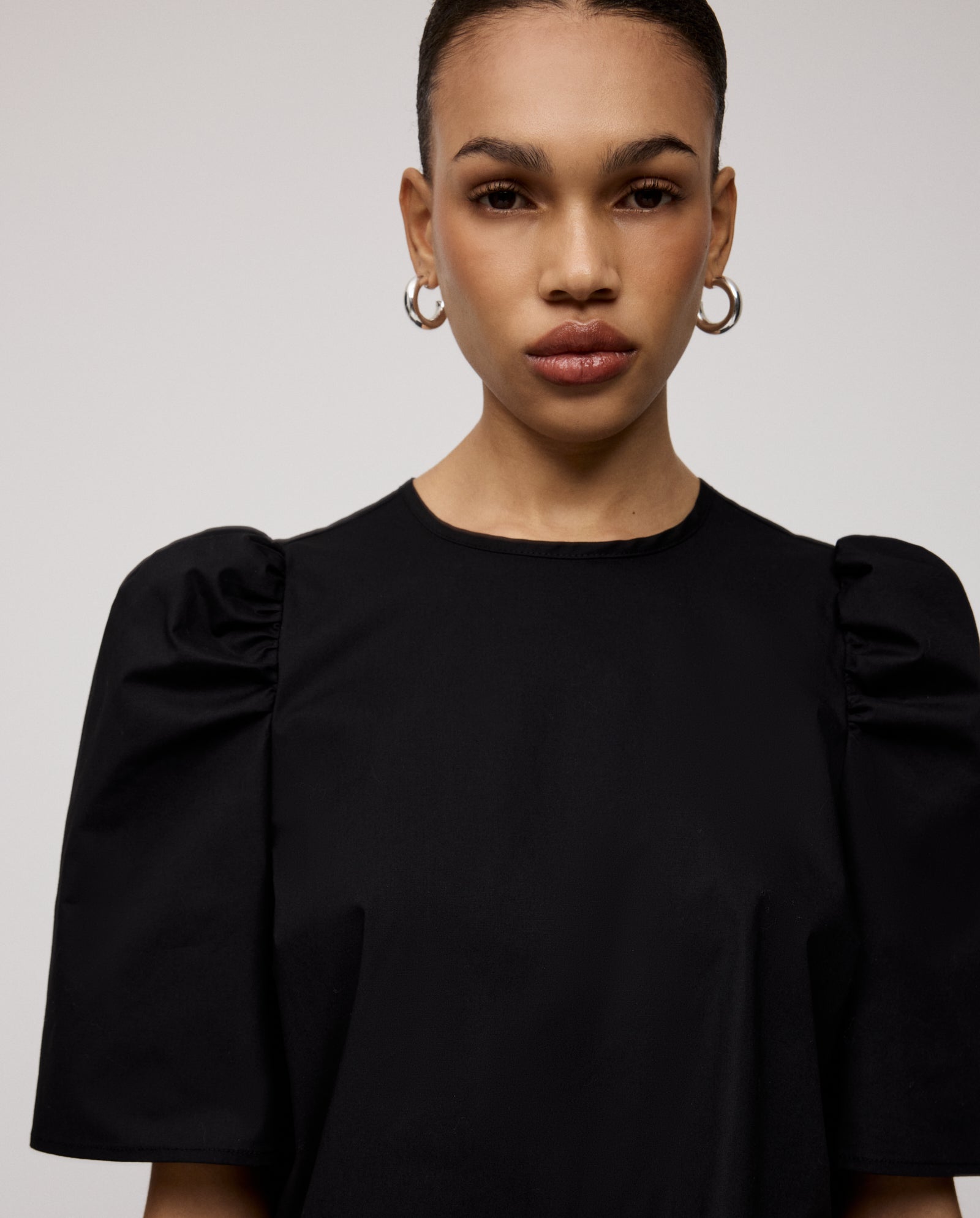 A woman wears the IVY OAK ELISABETH ANN Blouse—a black cropped cotton top with voluminous sleeves—paired with hoop earrings, looking straight at the camera with a neutral expression.