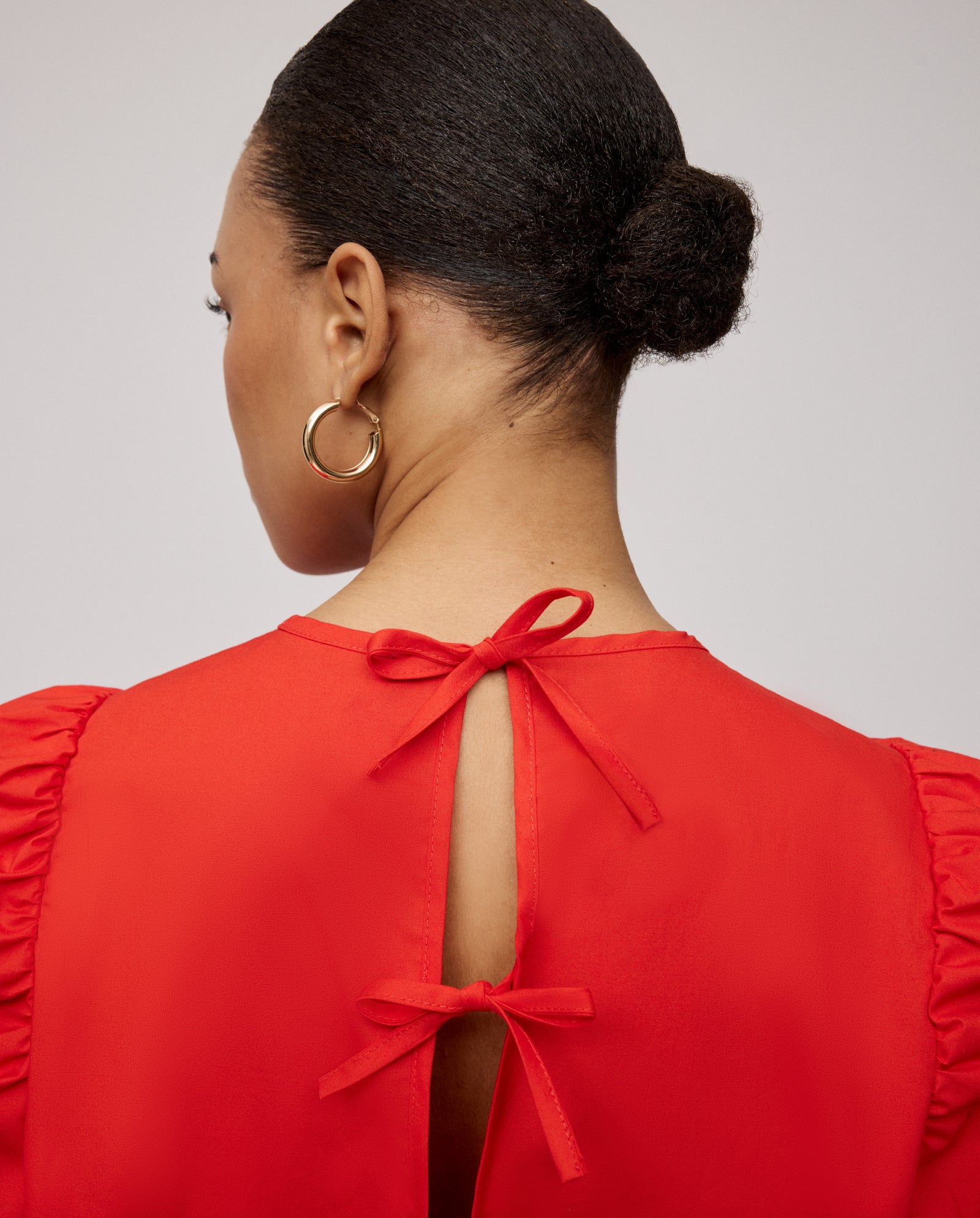 A woman with a neat bun wears gold hoop earrings and the IVY OAK ELISABETH ANN Blouse—a red cropped cotton top with voluminous sleeves and two bows tied at the open back.