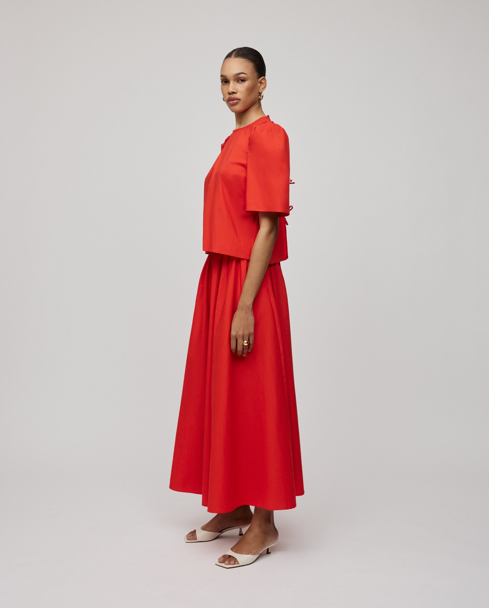 A woman wearing the IVY OAK ELISABETH ANN red cotton blouse with voluminous sleeves and a coordinated long skirt, standing sideways in white heels against a plain background.