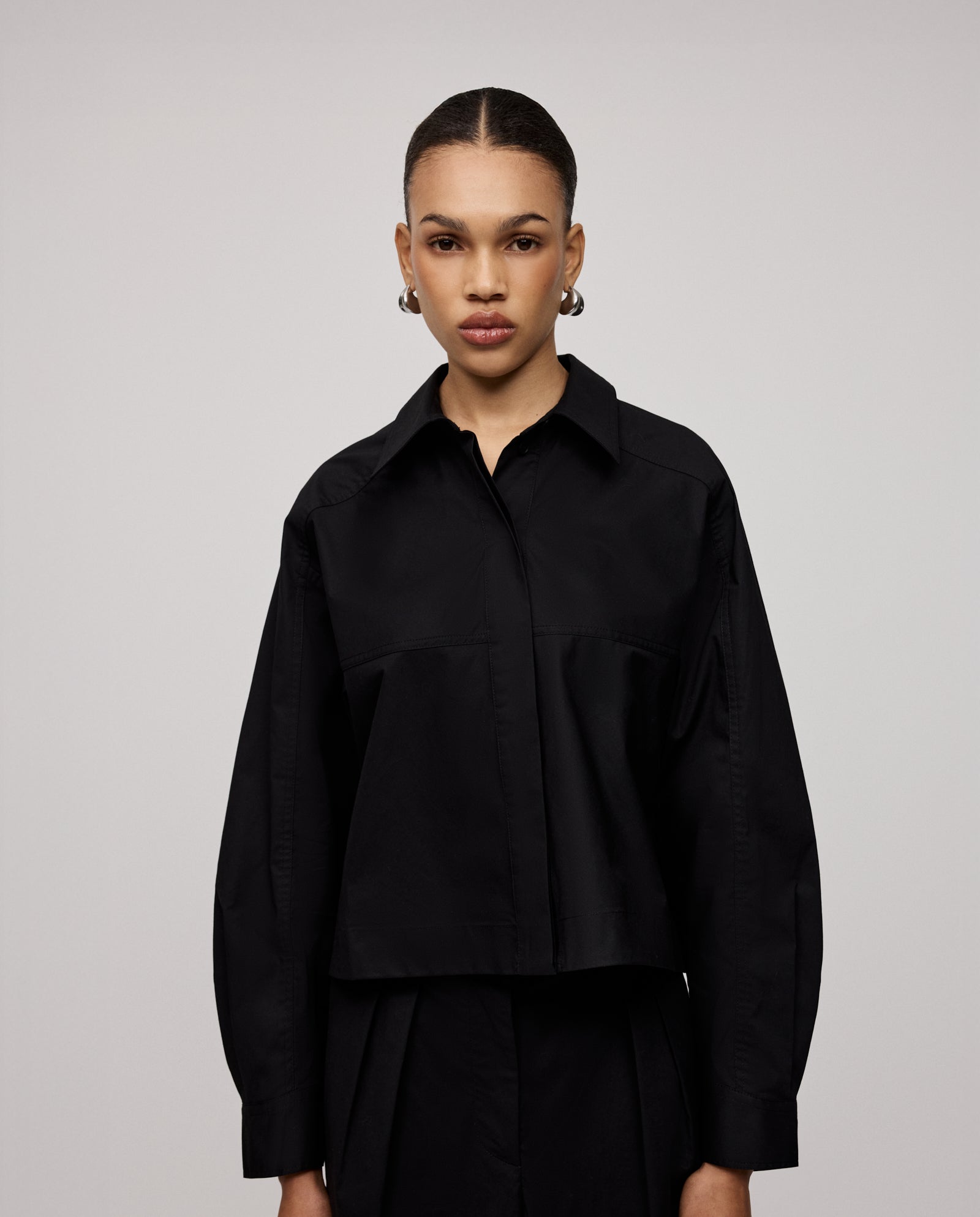 A woman wears the IVY OAK BRITTNEY Blouse, a black, boxy shirt with unique sleeve details, standing against a plain light background and looking directly at the camera.