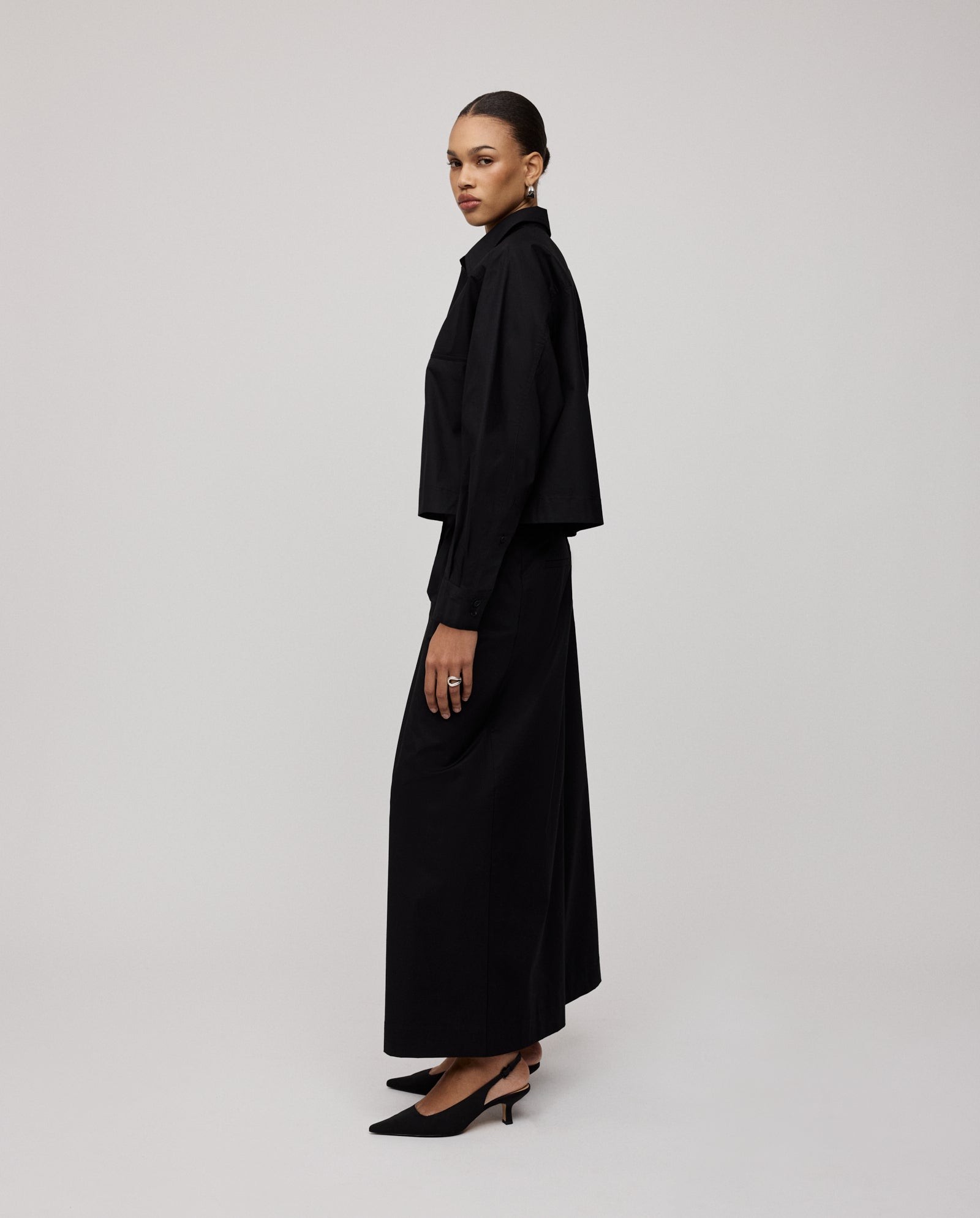 A woman wearing the IVY OAK BRITTNEY Blouse, paired with a black long skirt, boxy cropped jacket with unique sleeve detail, and heels, poses sideways against a plain light background.