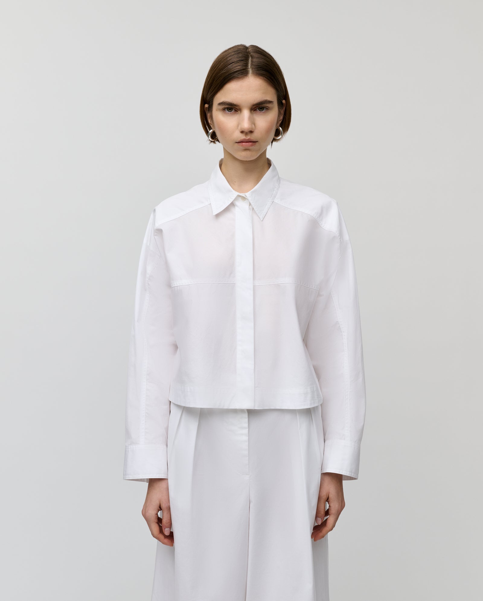 A person wears the IVY OAK BRITTNEY Blouse, a crisp white boxy shirt with a hidden button placket, paired with white pants against a plain light background.