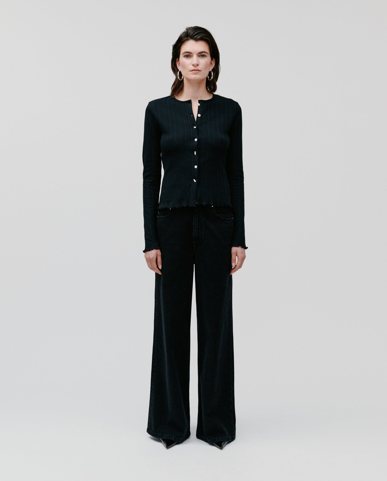 A woman stands against a plain light background, wearing the IVY OAK KAMILLE Cardigan in a classic fit with long sleeves, paired with wide-leg black pants.