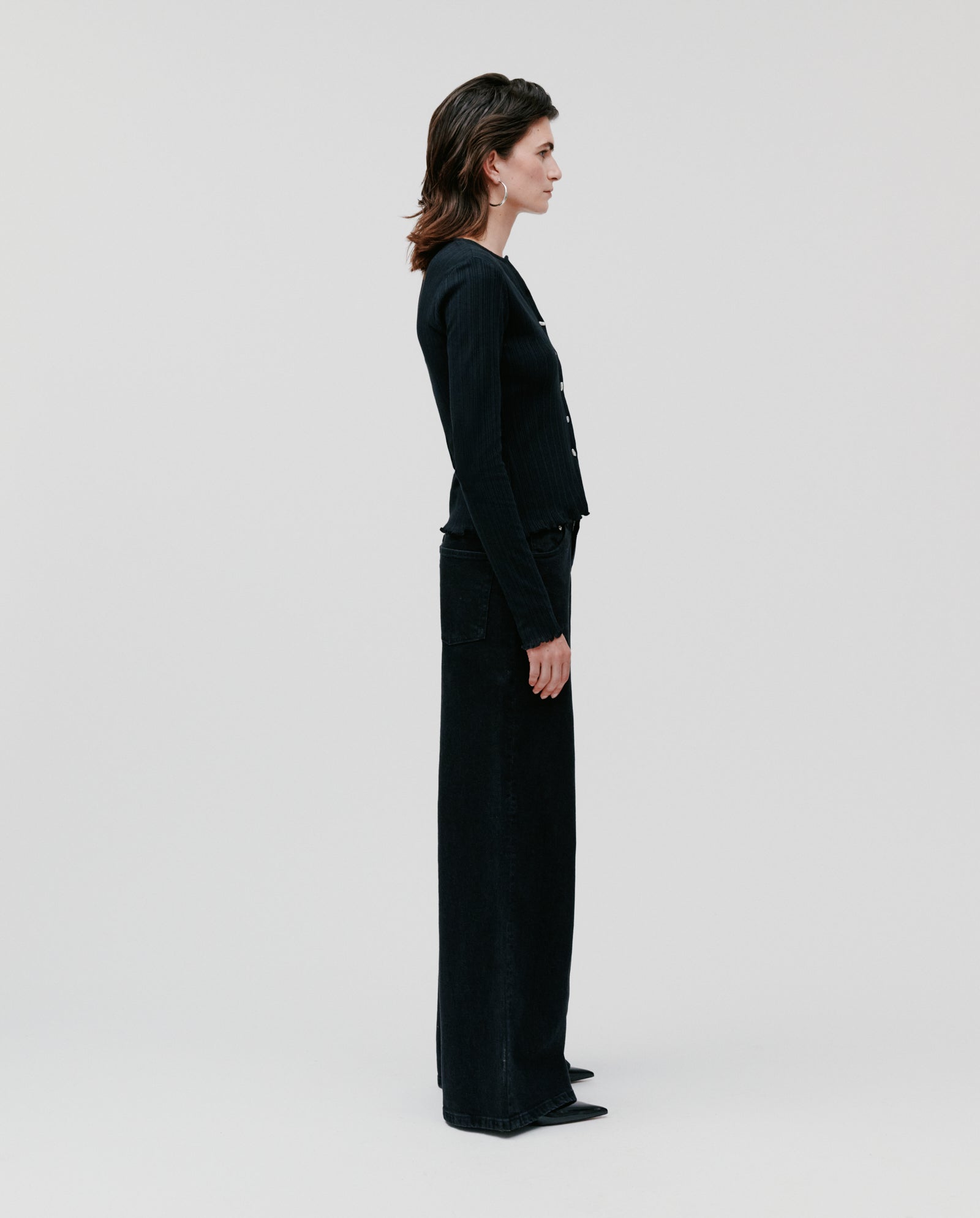 A person in profile wears the IVY OAK KAMILLE Cardigan, a classic-fit black cotton piece with long sleeves, paired with wide-leg black pants against a plain background.