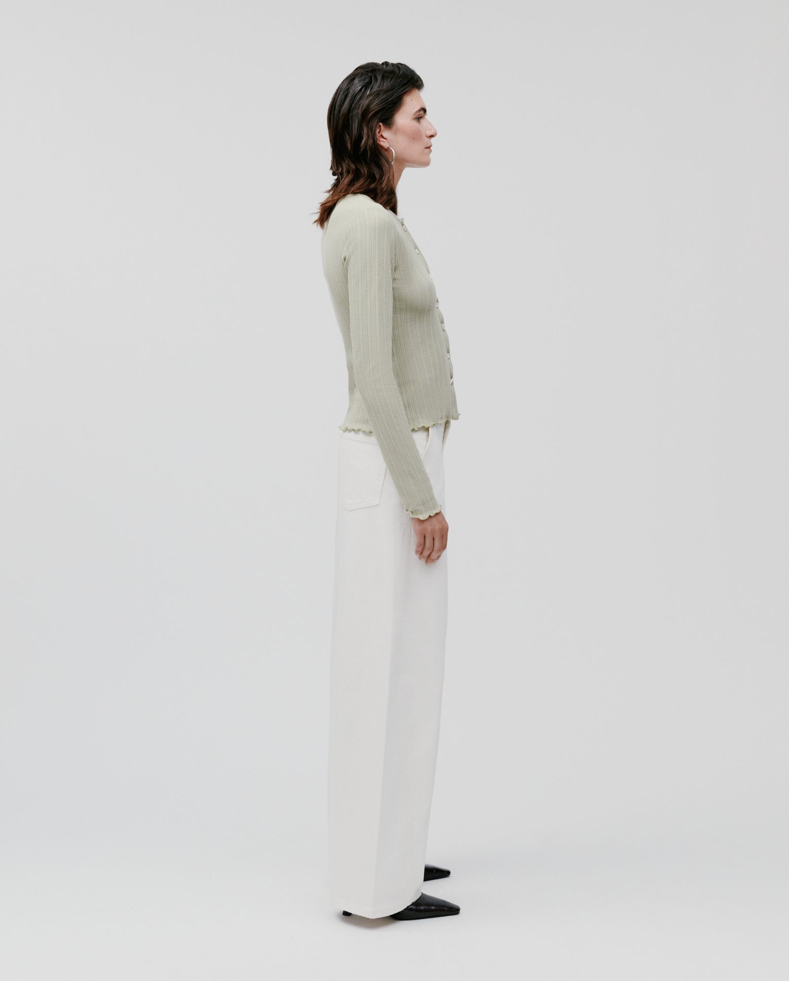 A person stands in profile against a plain background, wearing the IVY OAK KAMILLE Cardigan in classic fit cotton jersey paired with wide white pants.