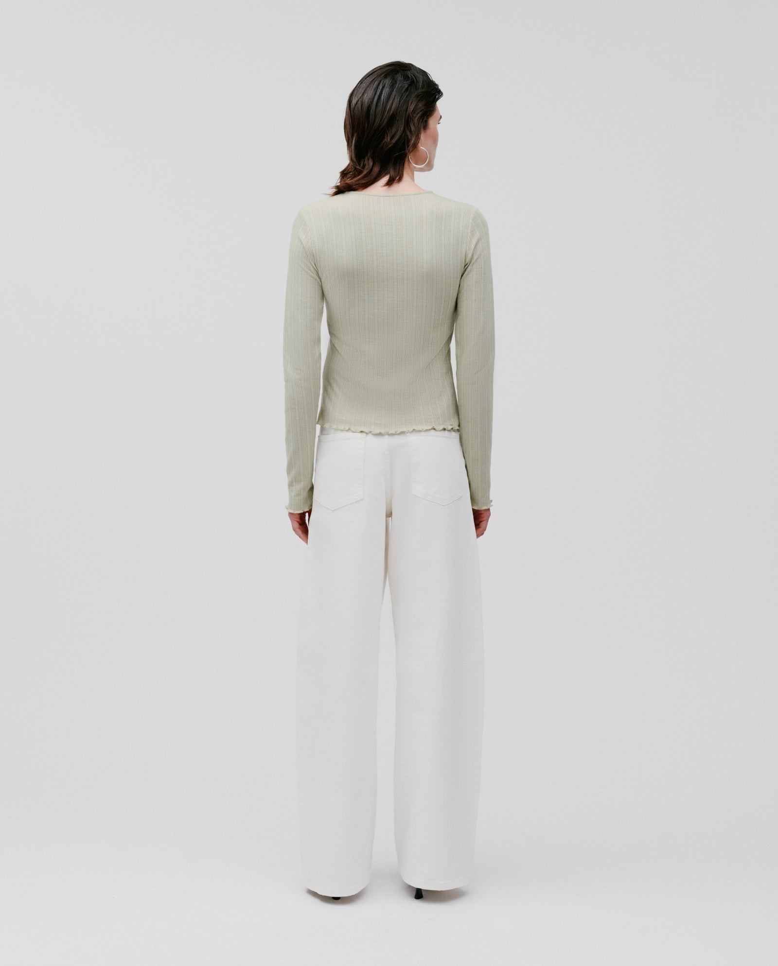 A woman stands facing away against a plain background, wearing the IVY OAK KAMILLE Cardigan in light green with wide-leg white pants.