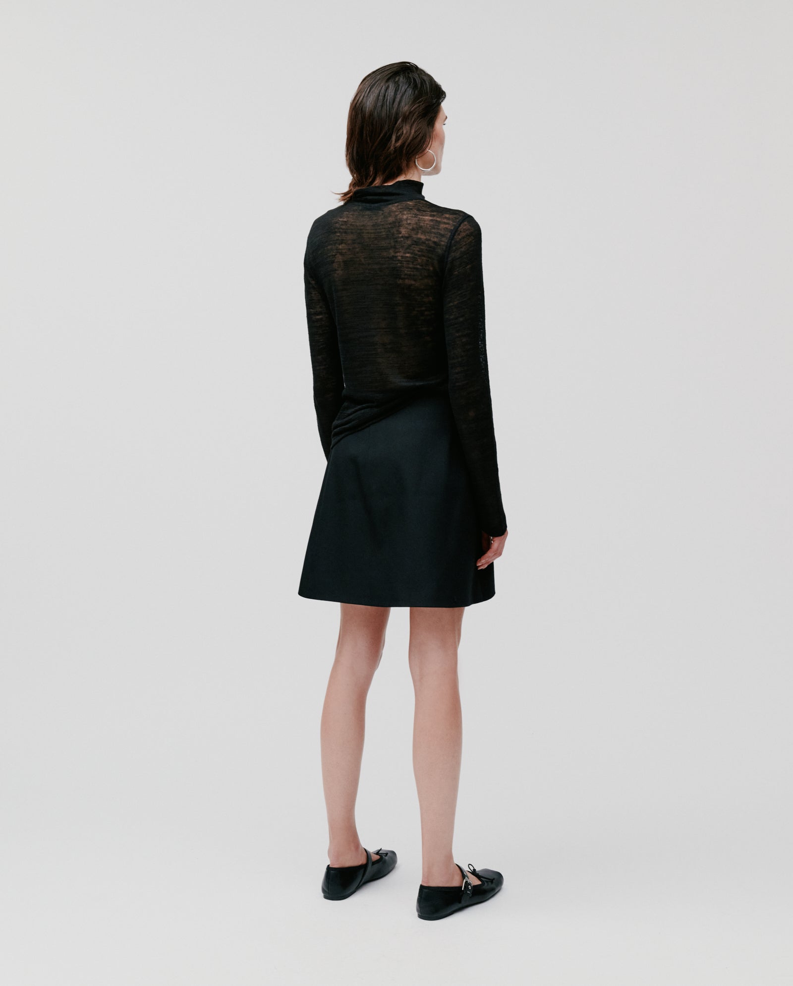 A woman stands facing away against a plain background, wearing the black sheer FELINE Top by IVY OAK, paired with a black skirt and black shoes.