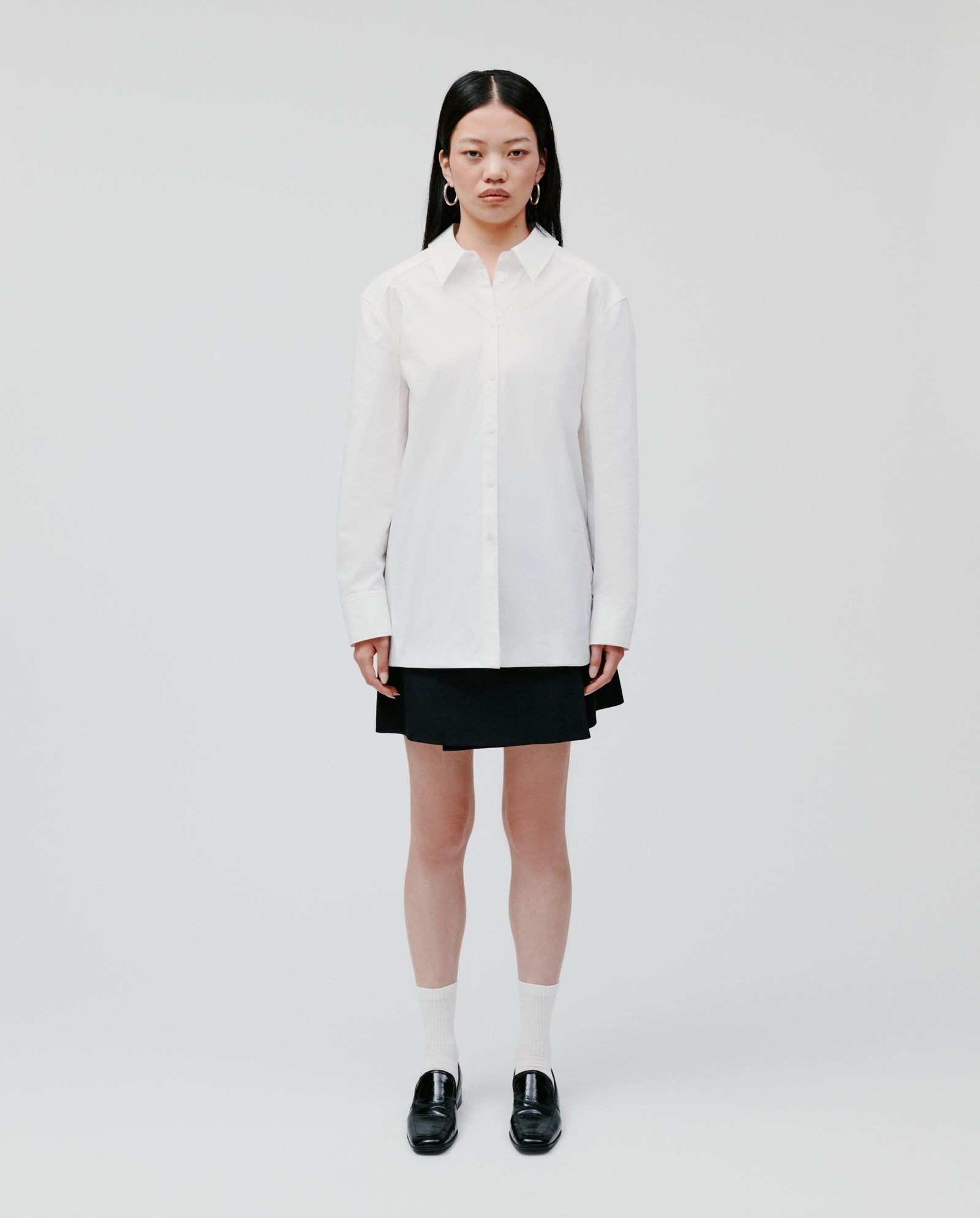 A woman stands against a white background wearing the IVY OAK EMORY Blouse, paired with a black skirt, white socks, and black shoes.