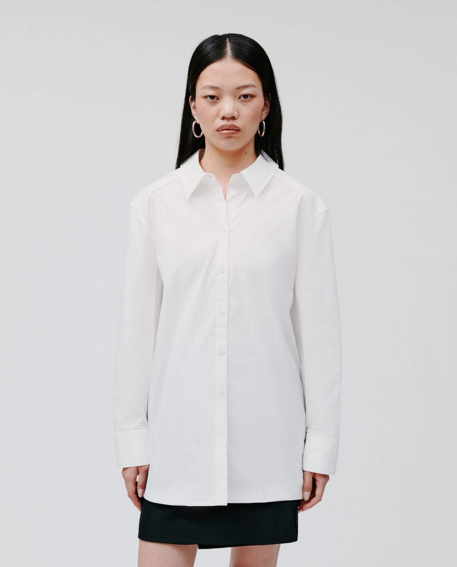 A woman with long dark hair wears the IVY OAK EMORY Blouse, a white cotton poplin top, paired with a black skirt and standing against a plain background.