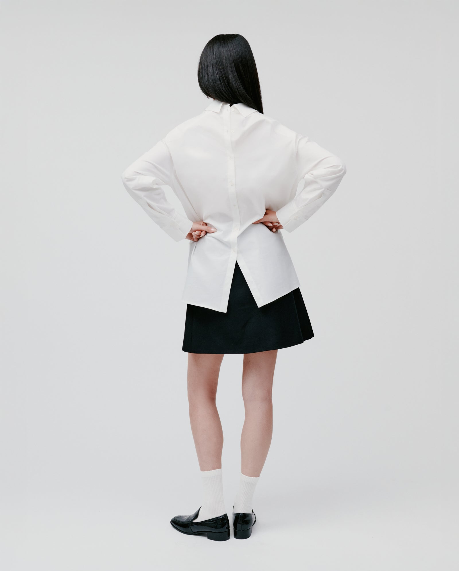 A woman stands with her back to the camera, wearing the IVY OAK EMORY Blouse in white cotton poplin, paired with a black skirt, socks, and loafers.