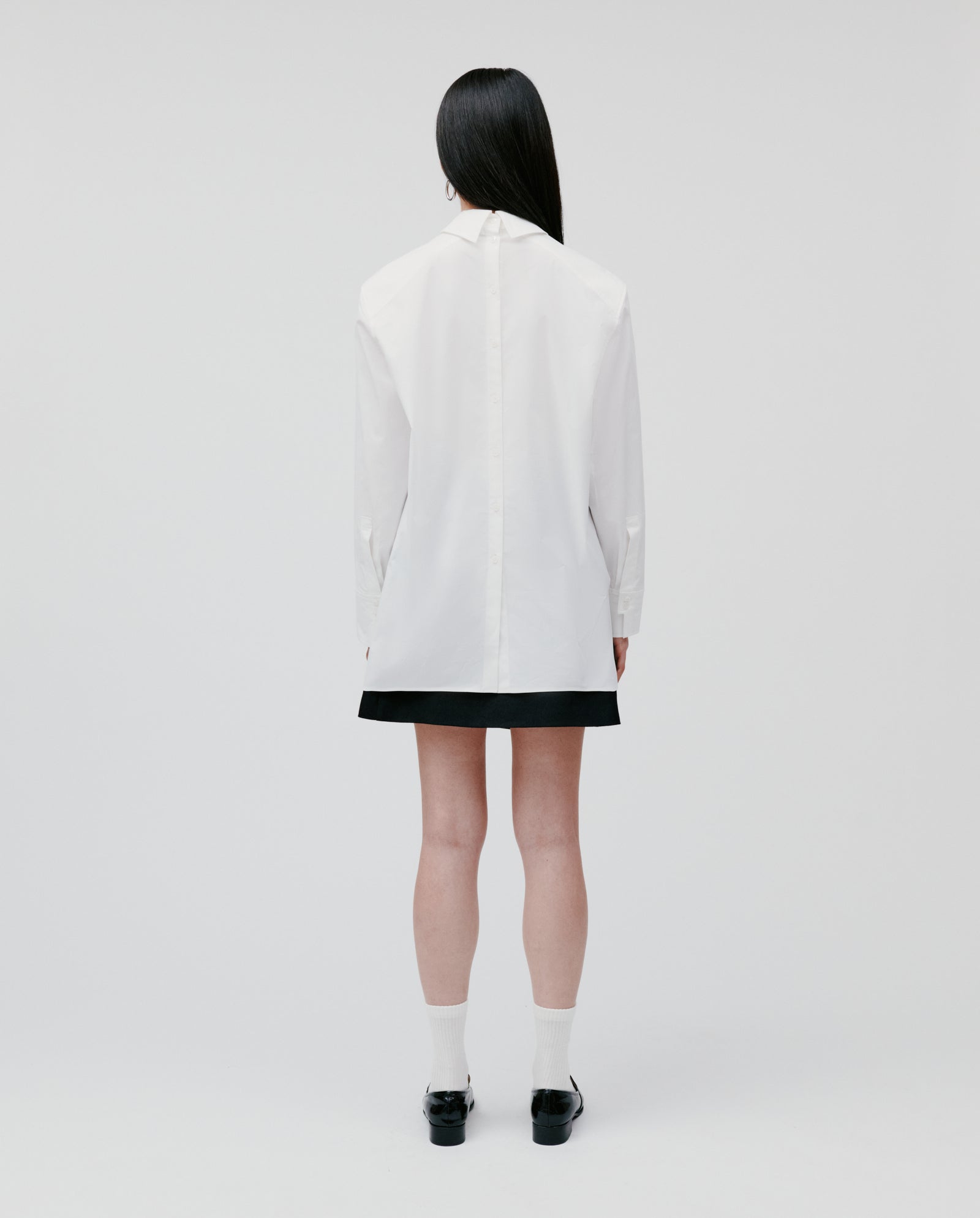 A woman stands facing away on a plain background, wearing the IVY OAK EMORY Blouse in white cotton popline, styled with a black skirt, white socks, and black shoes.