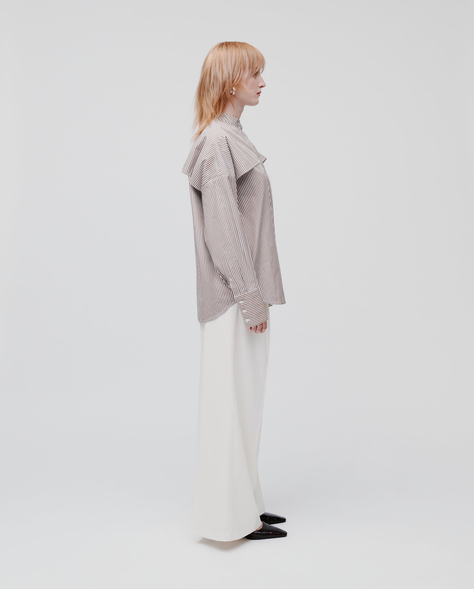 A person in profile wears the IVY OAK EVE Blouse—an oversize striped shirt with button closure—paired with white wide-leg pants and black shoes, set against a plain background.
