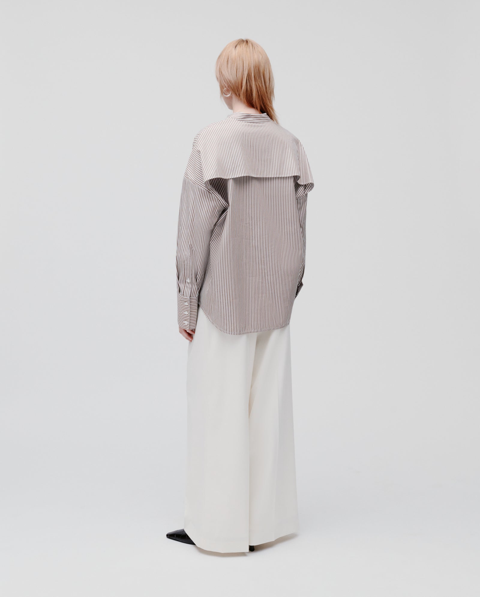 A person stands backwards in an IVY OAK EVE Blouse—an oversize striped shirt with button closure—paired with wide white pants against a plain light background.
