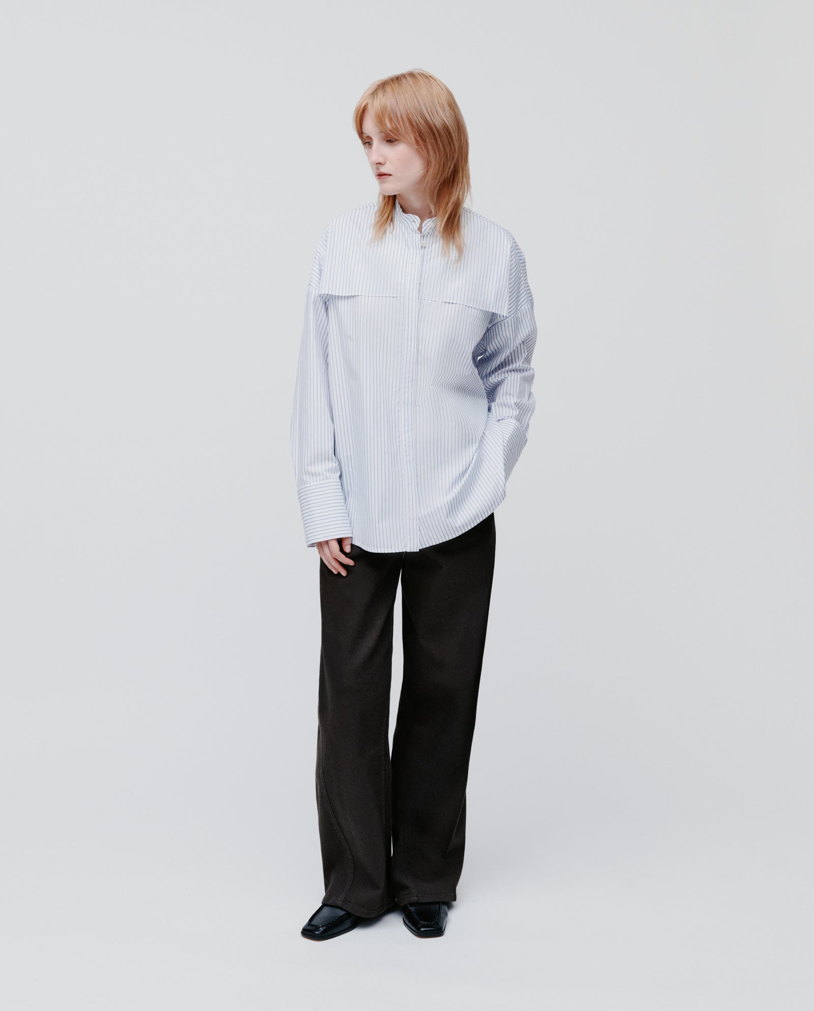 Model in the IVY OAK EVE Blouse—an oversize striped shirt with button closure—paired with wide-leg black pants, standing against a plain white background.