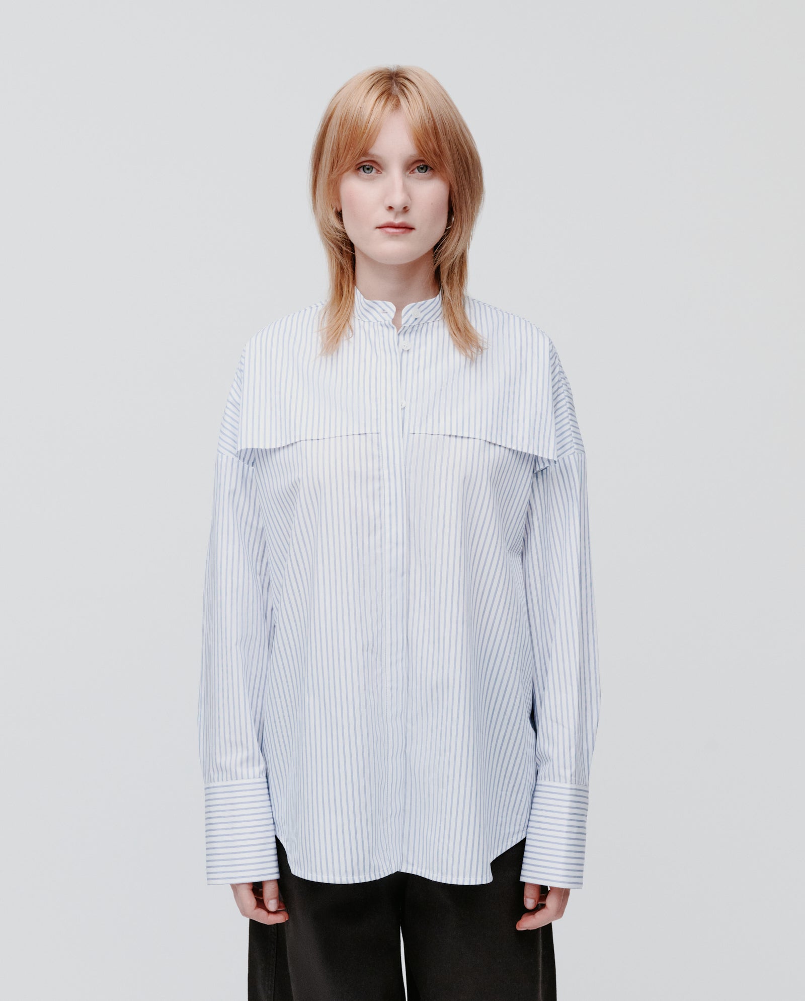 A person stands facing forward against a plain background, wearing the IVY OAK EVE Blouse—an oversize striped shirt with button closure—paired with black pants.