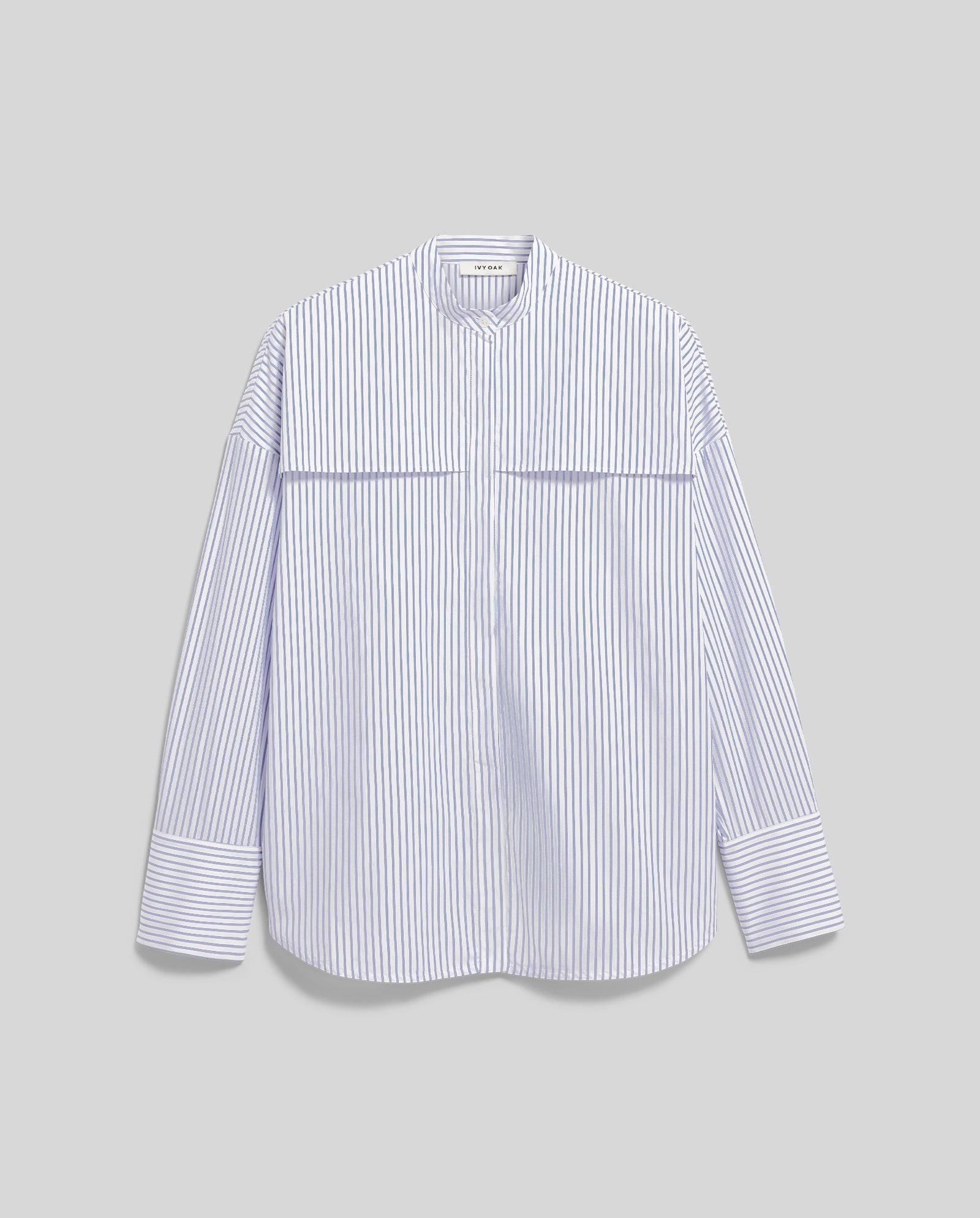 The EVE Blouse by IVY OAK is a white, long-sleeve oversized shirt with thin blue vertical stripes, a band collar, wide cuffs, and button closure, shown on a gray background.