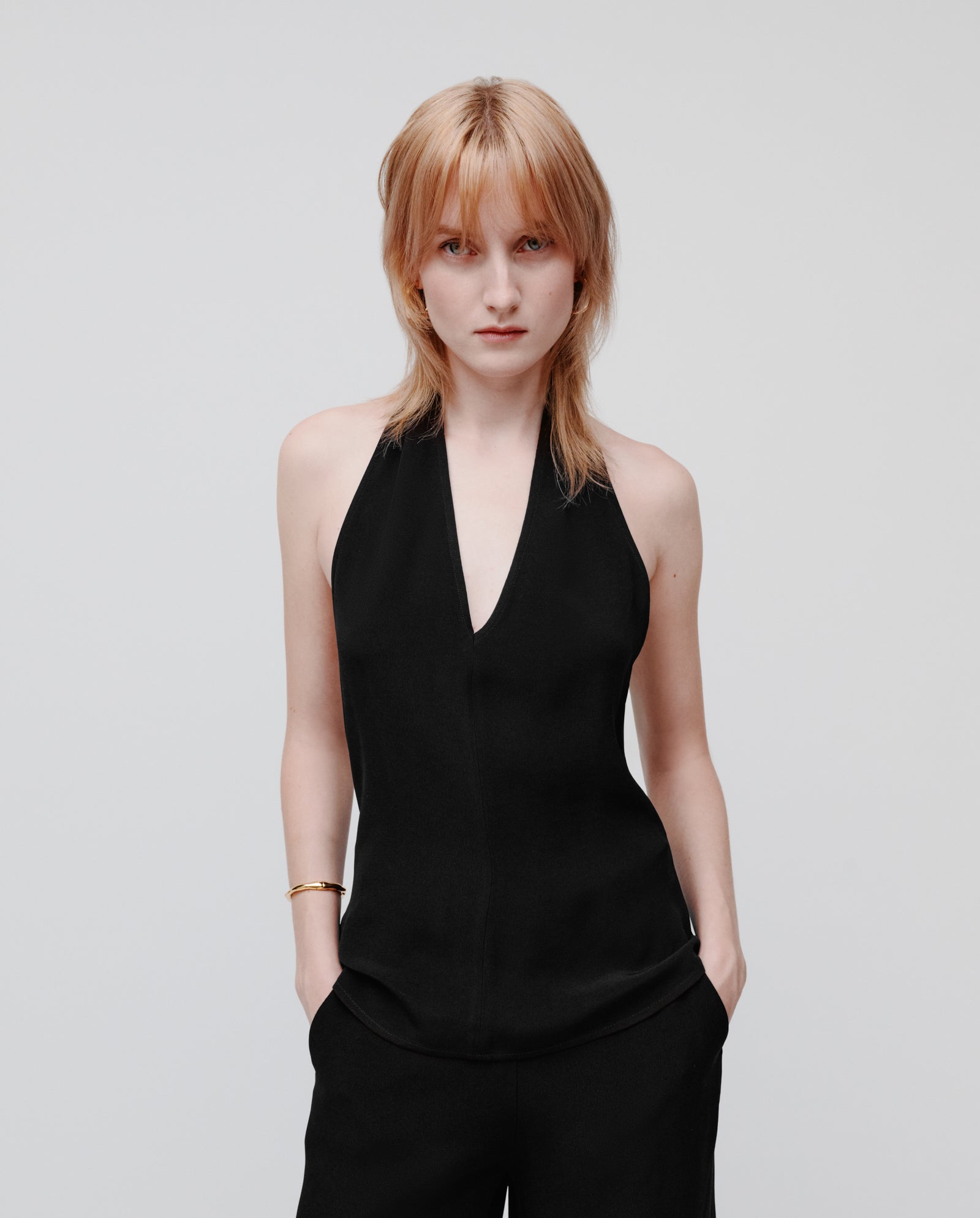 A light-haired person wears the IVY OAK TANSY Top, a black crepe neckholder style, with matching pants, standing against a plain white background with hands in pockets.