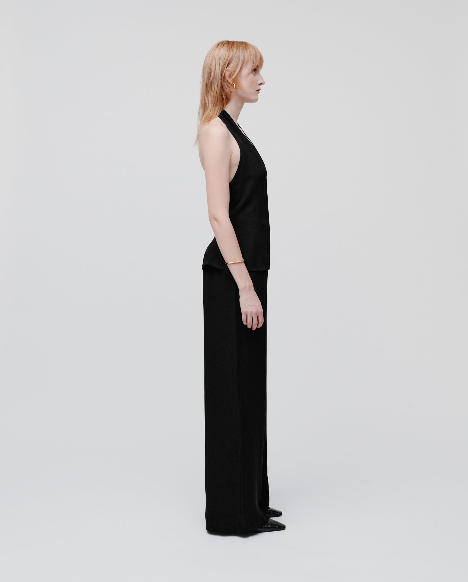 A woman with light hair stands in profile, wearing the IVY OAK TANSY Top—a sleeveless neckholder style—paired with elegant wide-leg black crepe pants.