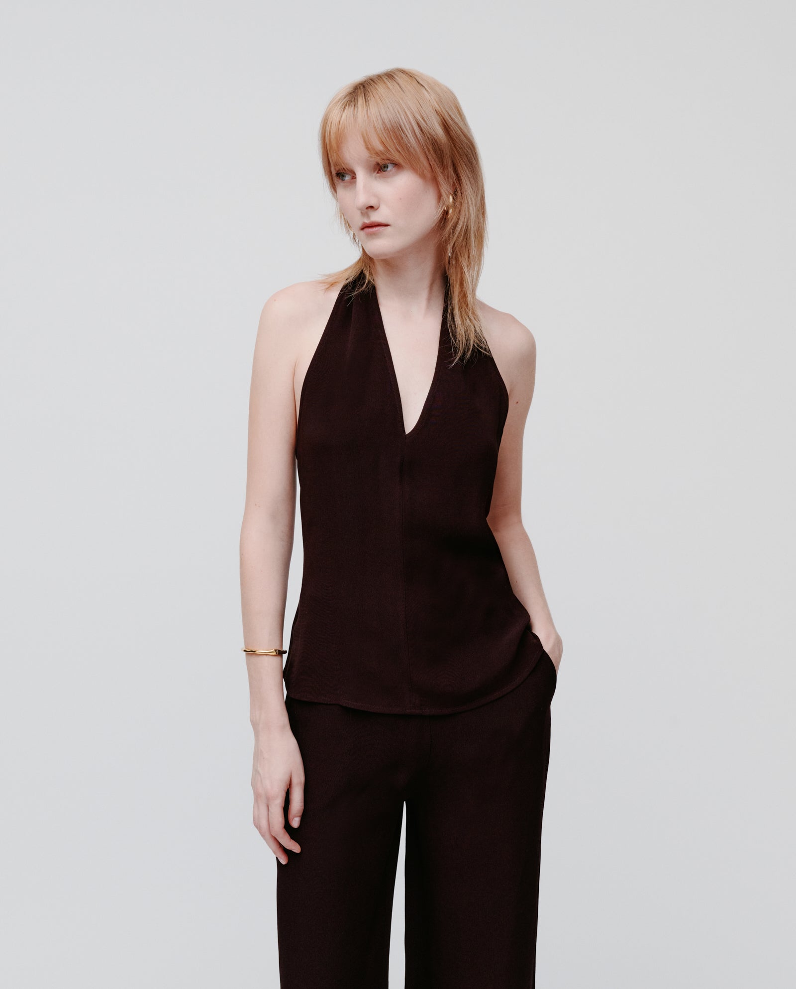 A woman wears the IVY OAK TANSY Top, a sleeveless black neckholder, paired with black pants. She stands with one hand in her pocket against a plain background, the chic crepe fabric adding elegance to her look.