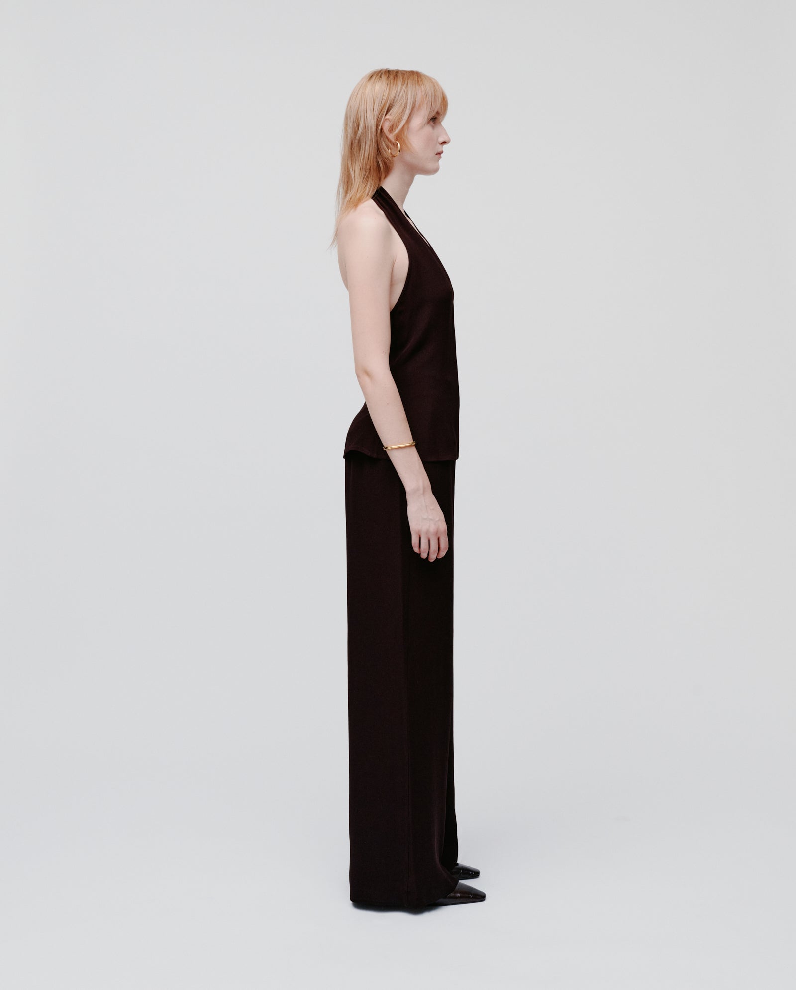 A woman stands in profile against a plain background, wearing the IVY OAK TANSY Top—a sleeveless black crepe neckholder—paired with matching black pants featuring a zipper side seam.