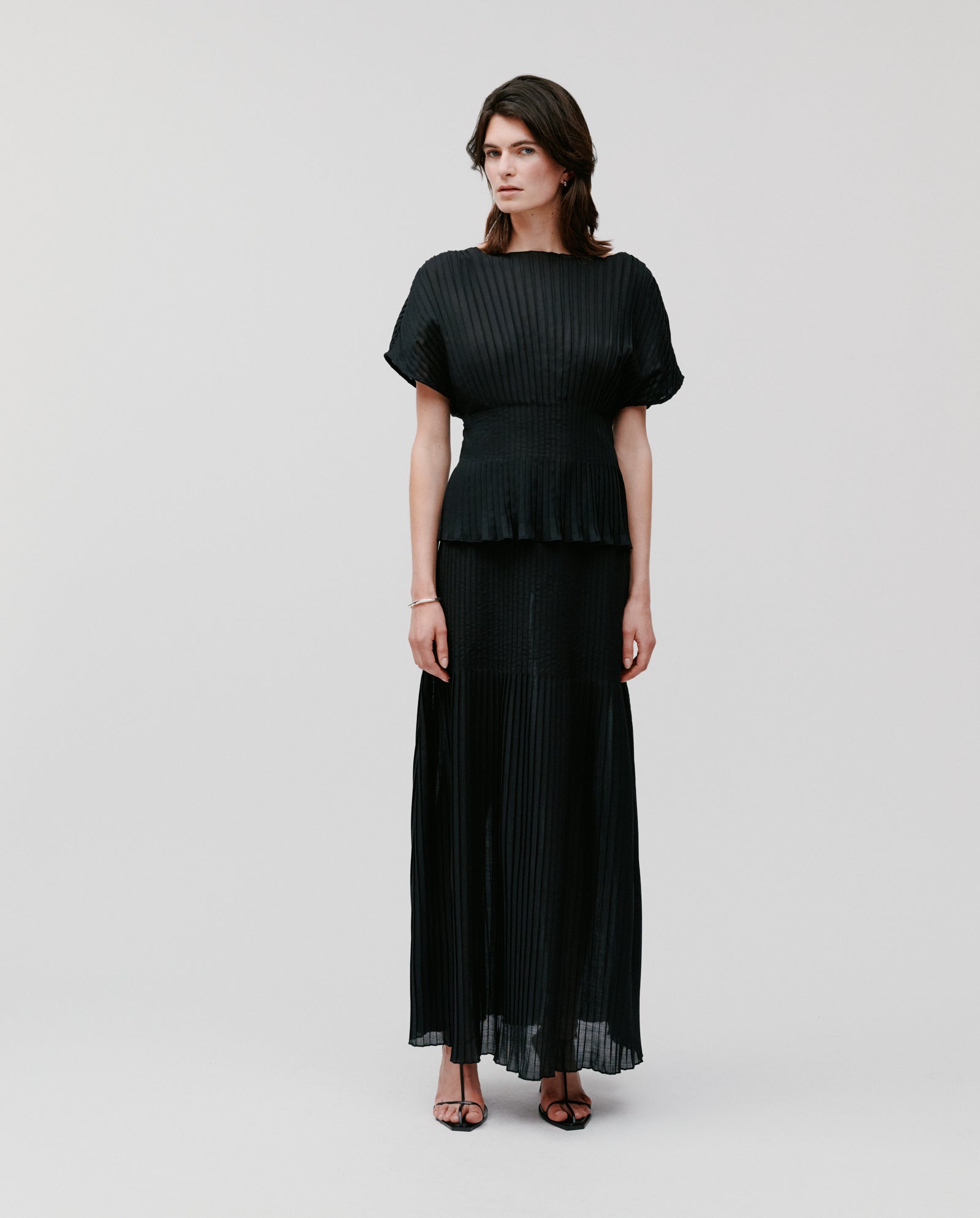 A woman wears the IVY OAK TAYE Top in black plissé fabric with a long skirt, standing against a plain light background.