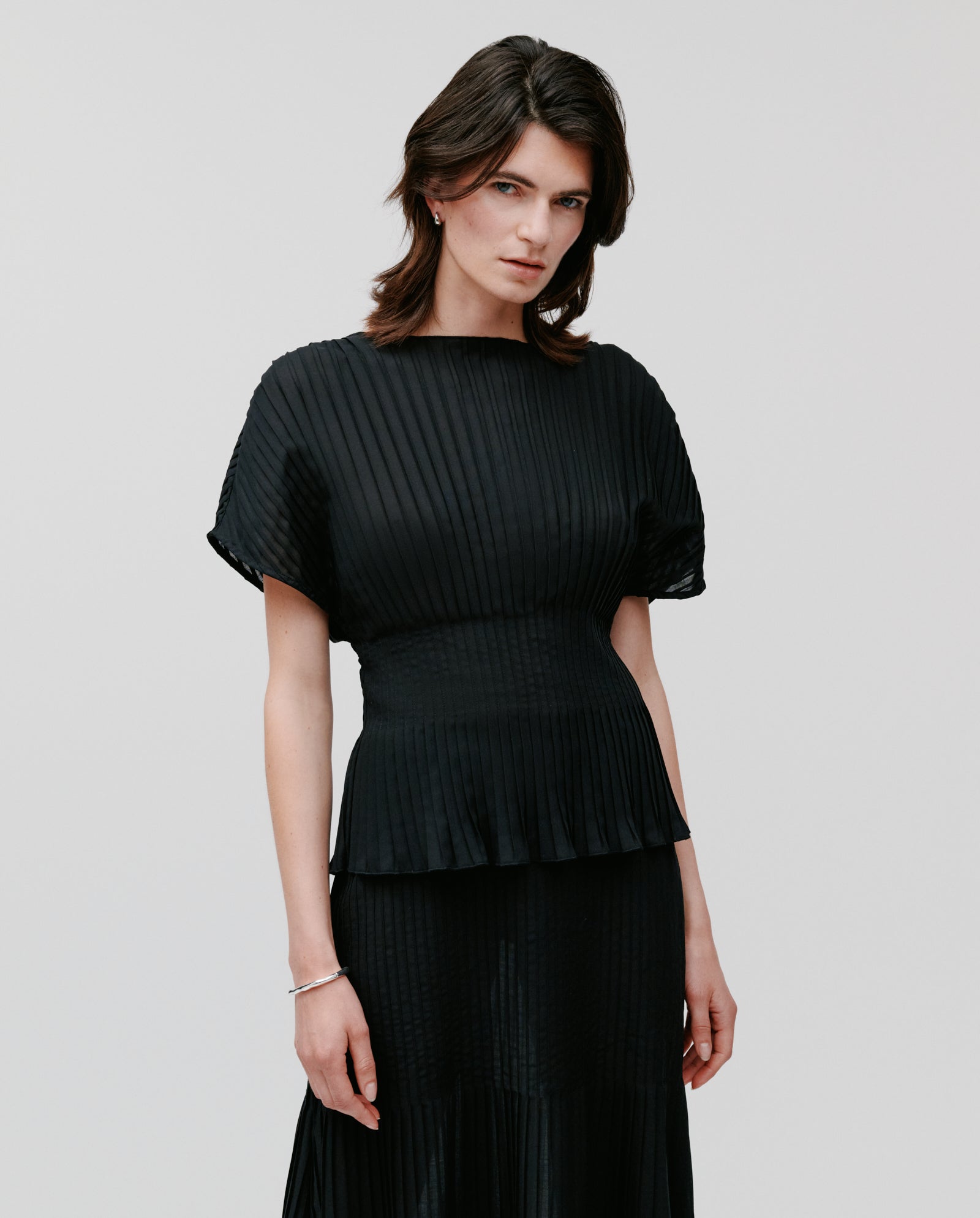 A woman wears the IVY OAK TAYE Top, featuring pleats and kimono sleeves, standing against a plain light gray background.