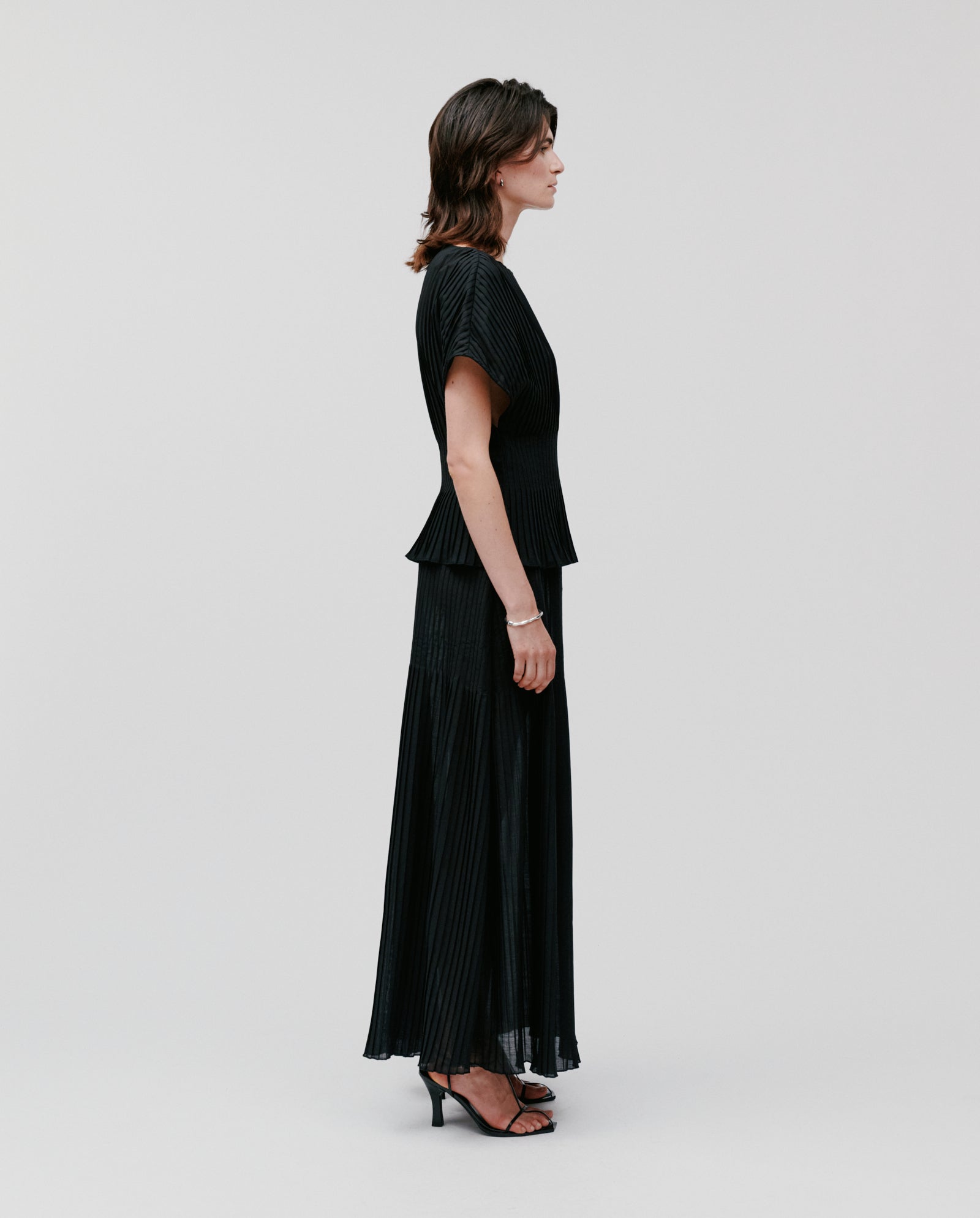 A woman in profile wears the IVY OAK TAYE Top in black plissé with kimono sleeves, paired with a long skirt and heels, set against a plain background.
