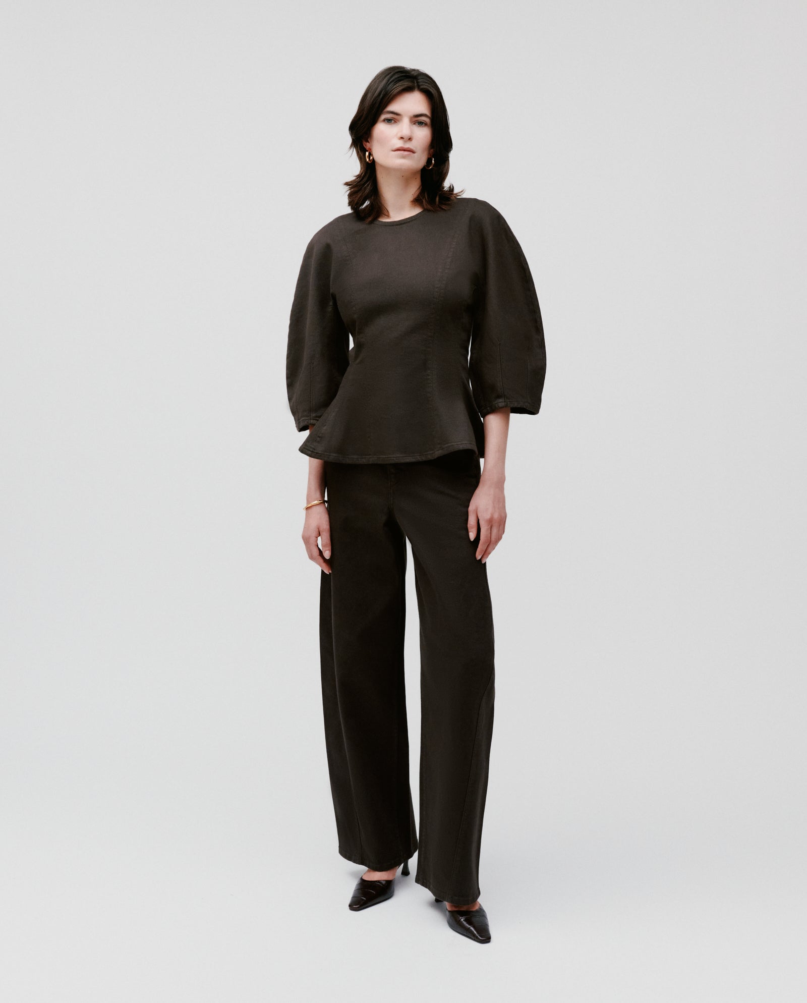 A woman stands against a plain background wearing the IVY OAK ELMA Blouse with bell-shaped sleeves, paired with wide-legged dark pants.