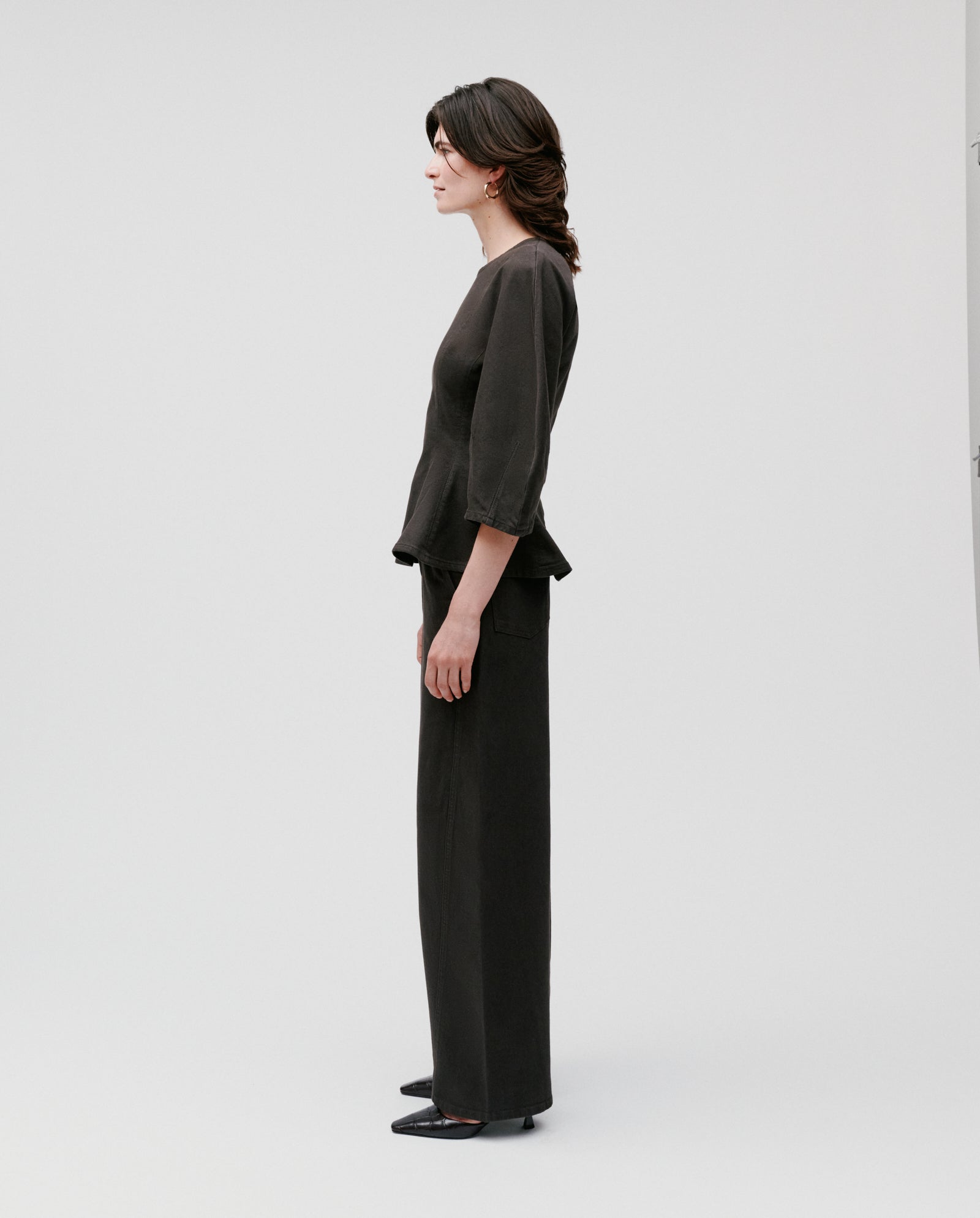 A woman stands in profile against a plain background, wearing the IVY OAK ELMA Blouse—a loose black top with bell sleeves—paired with wide-leg black pants.