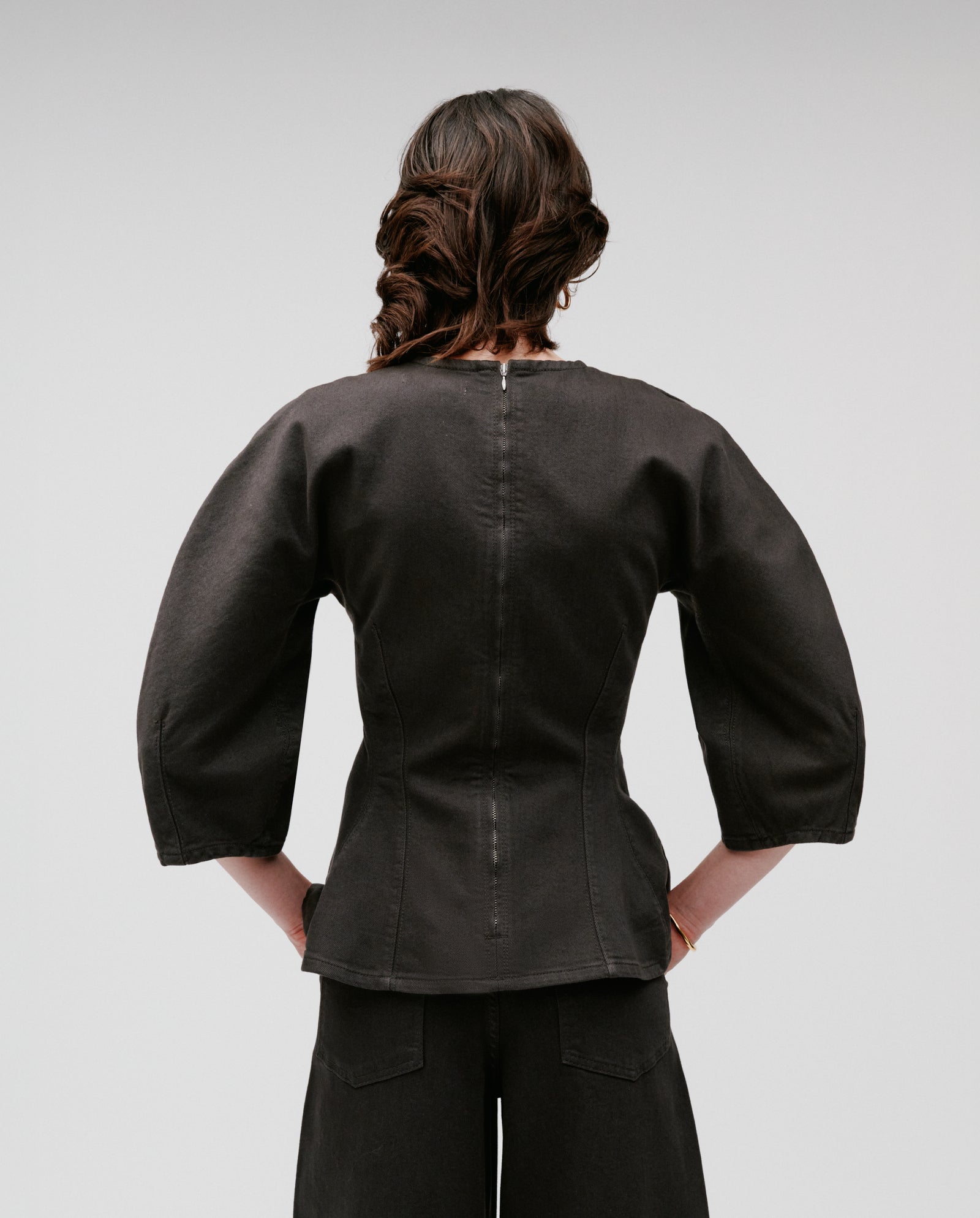 A woman with wavy hair stands facing away, wearing the IVY OAK ELMA Blouse in dark denim with bell sleeves and matching pants.