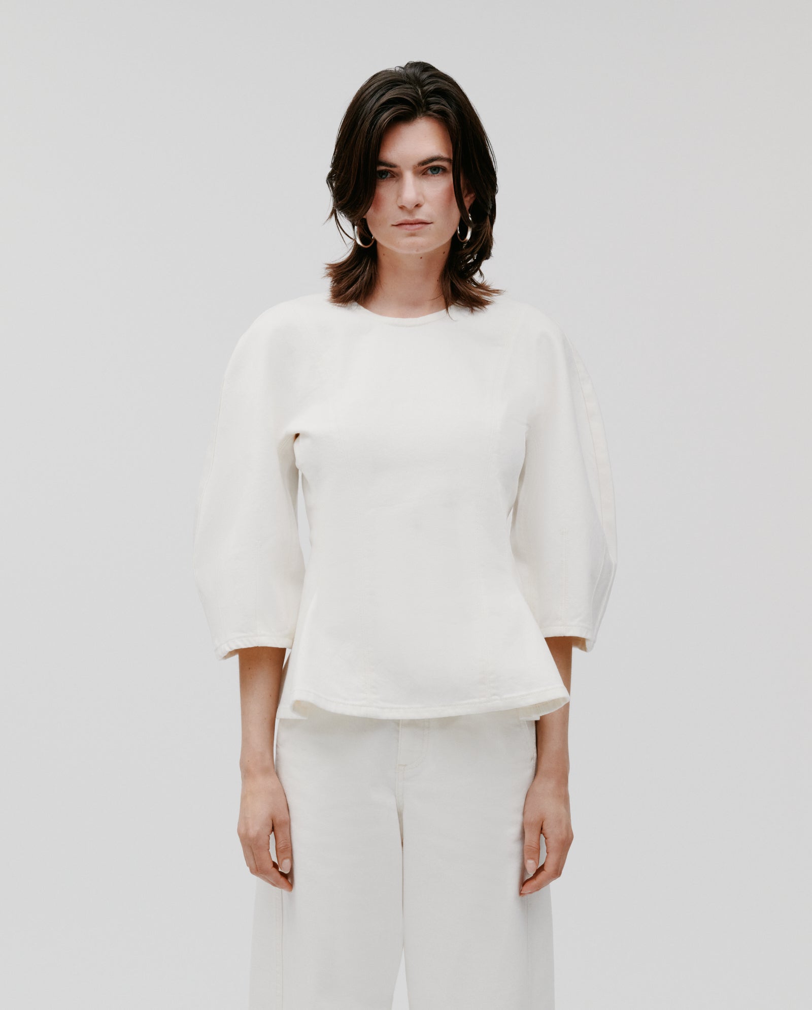 Model with shoulder-length dark hair wears the IVY OAK ELMA Blouse, featuring a loose fit and wide-shoulder silhouette, paired with white pants against a plain background.