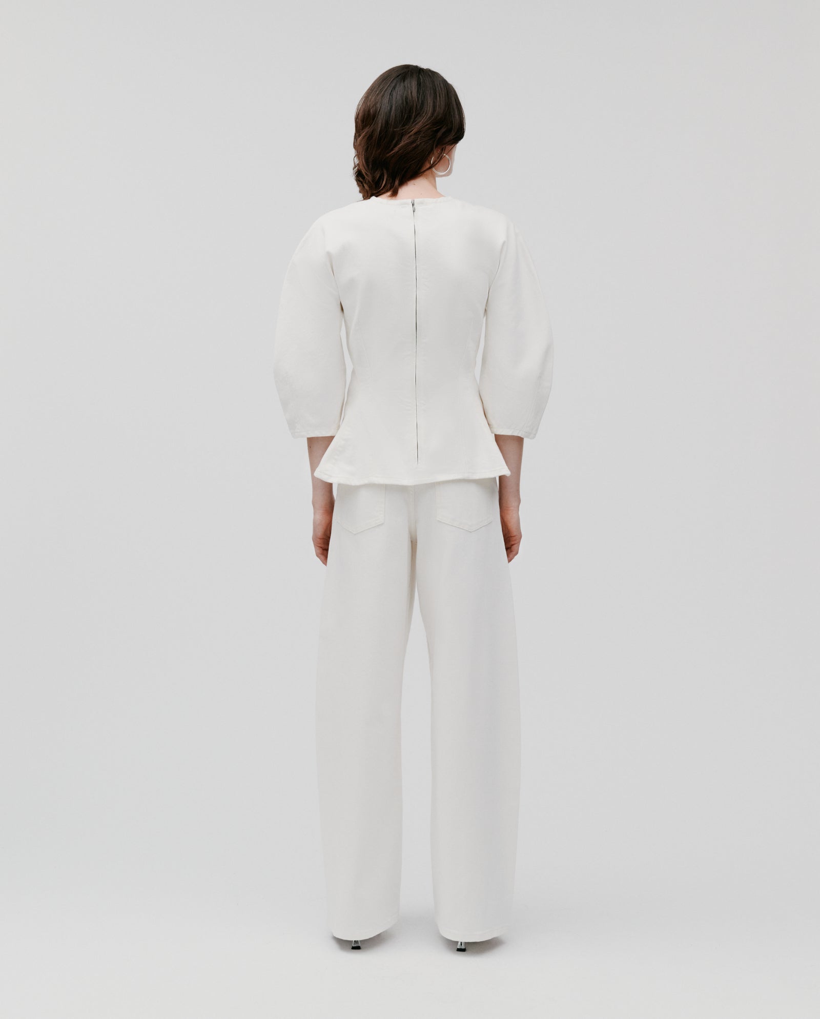 A person stands facing away on a plain background, wearing the IVY OAK ELMA Blouse—a white, wide-shoulder top with 3/4 sleeves—paired with wide-leg pants for a subtle peplum effect.