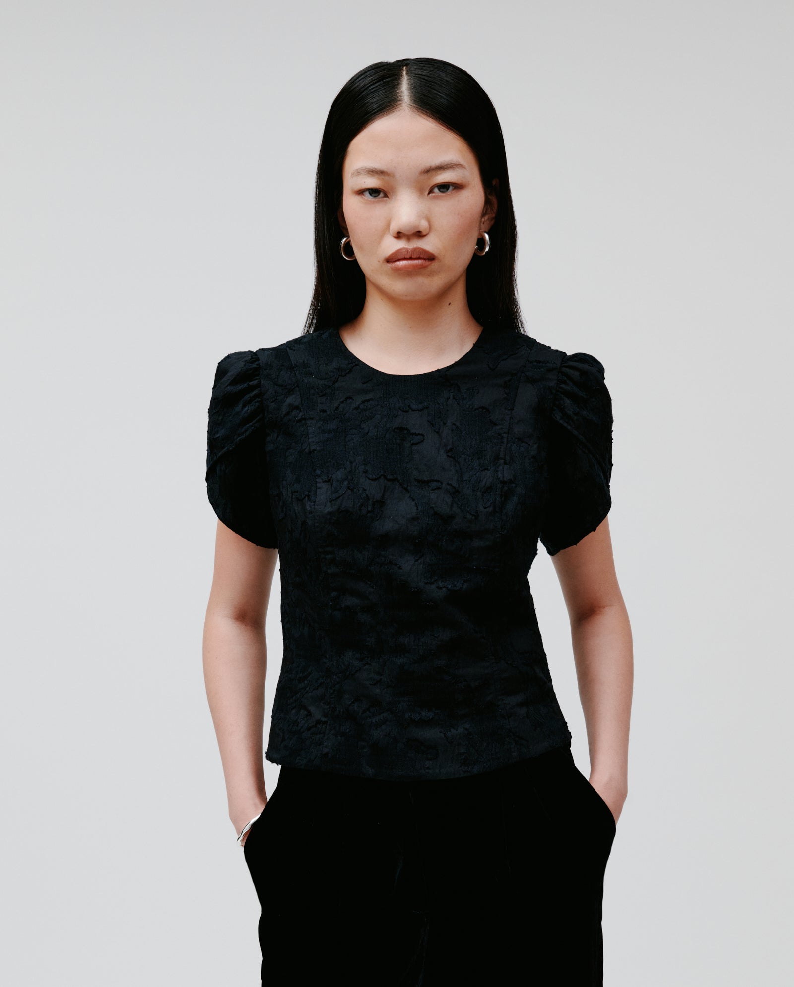 A woman with long dark hair models the IVY OAK ELODIE Blouse, a black fil coupé design featuring voluminous sleeves, as she stands with her hands in her pockets against a plain background.