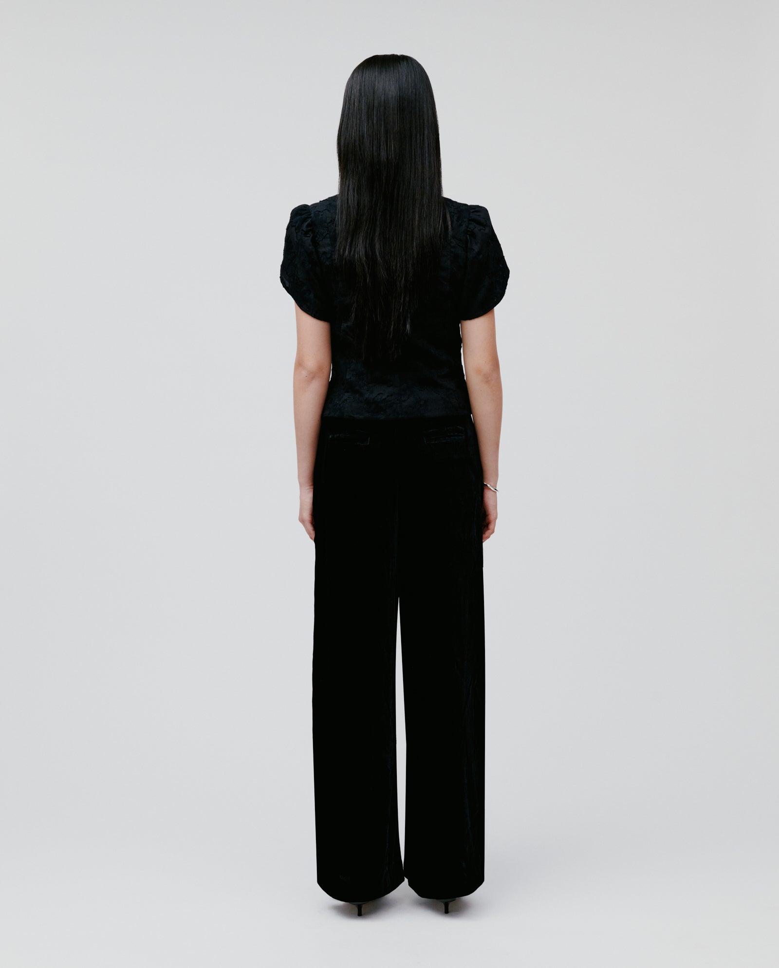 A woman with long dark hair faces away, wearing the IVY OAK ELODIE Blouse with voluminous sleeves and wide-leg black pants, creating a modern romantic silhouette.
