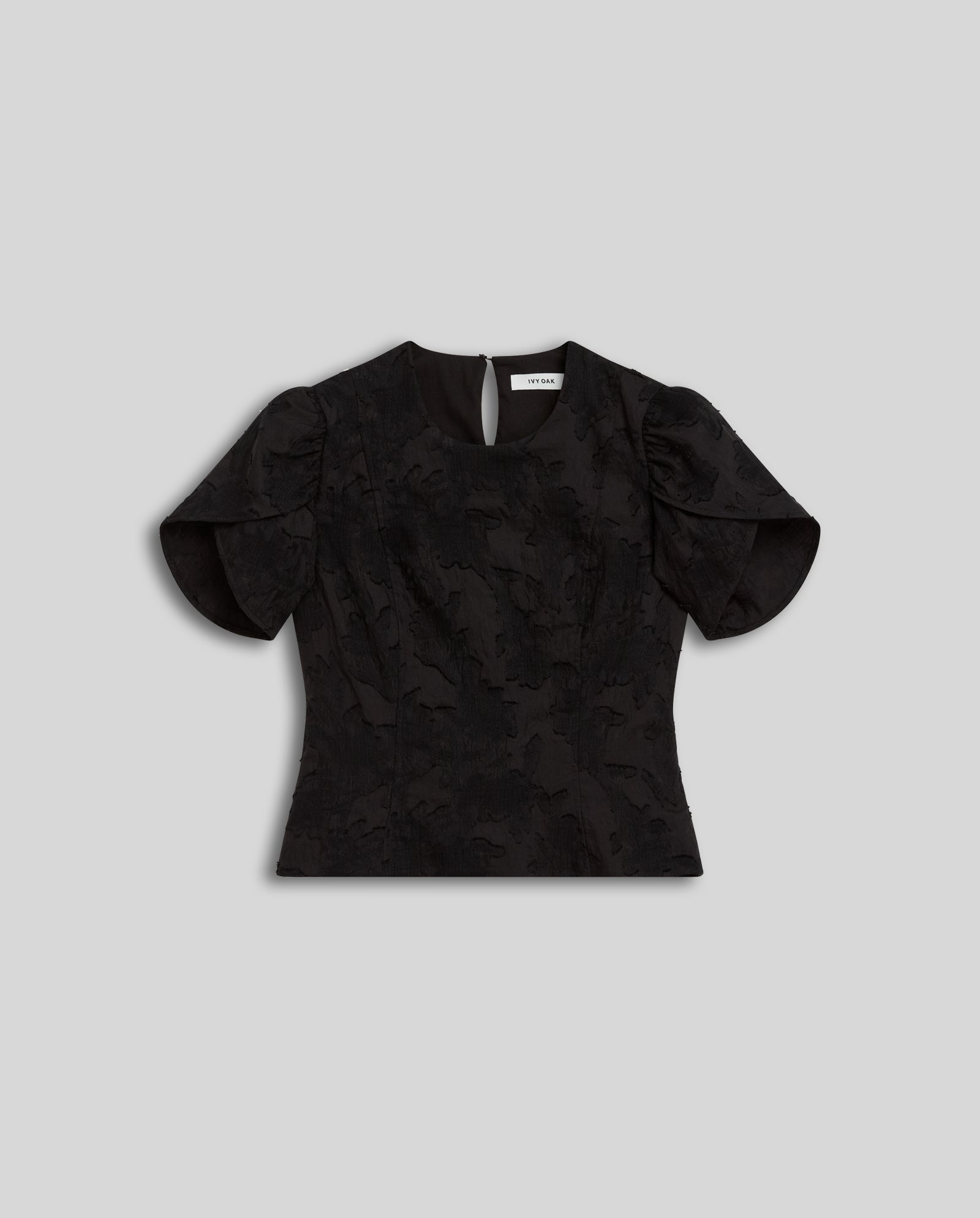 The ELODIE Blouse by IVY OAK is a short-sleeve black fil coupé top with voluminous puffed sleeves and a round neckline, offering a modern romantic silhouette, shown on a grey background.