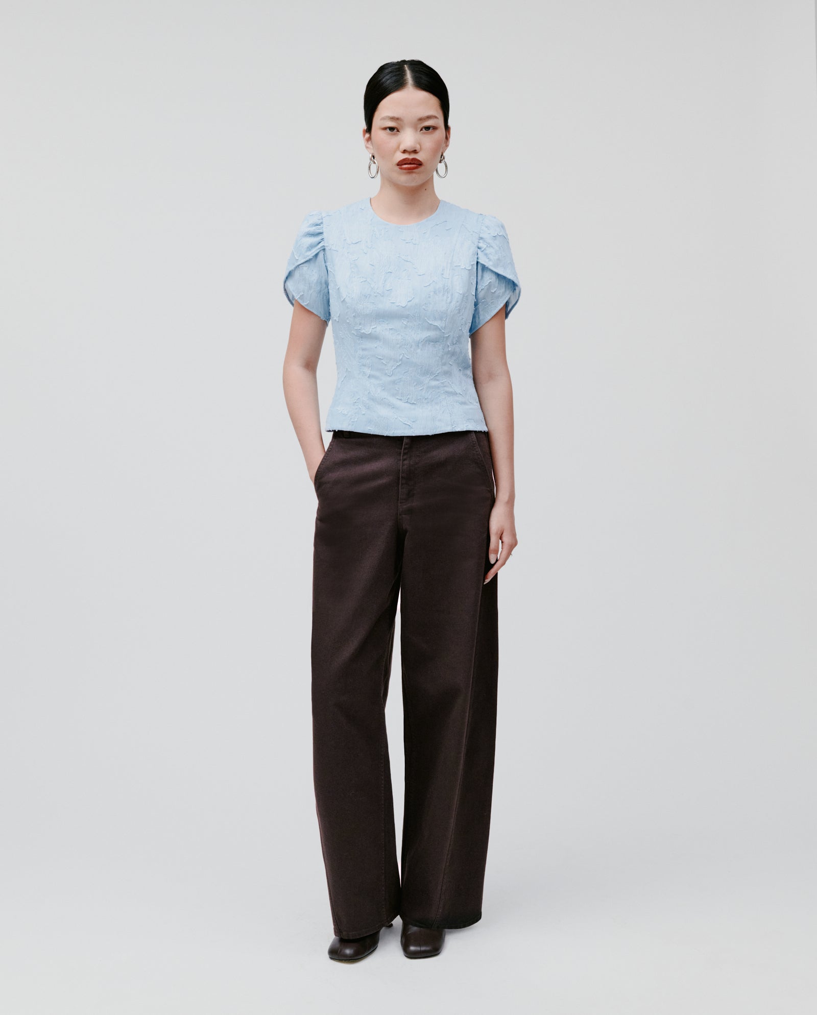 A woman wears the IVY OAK ELODIE Blouse in light blue, featuring voluminous sleeves, paired with wide-leg dark pants as she stands against a plain background.