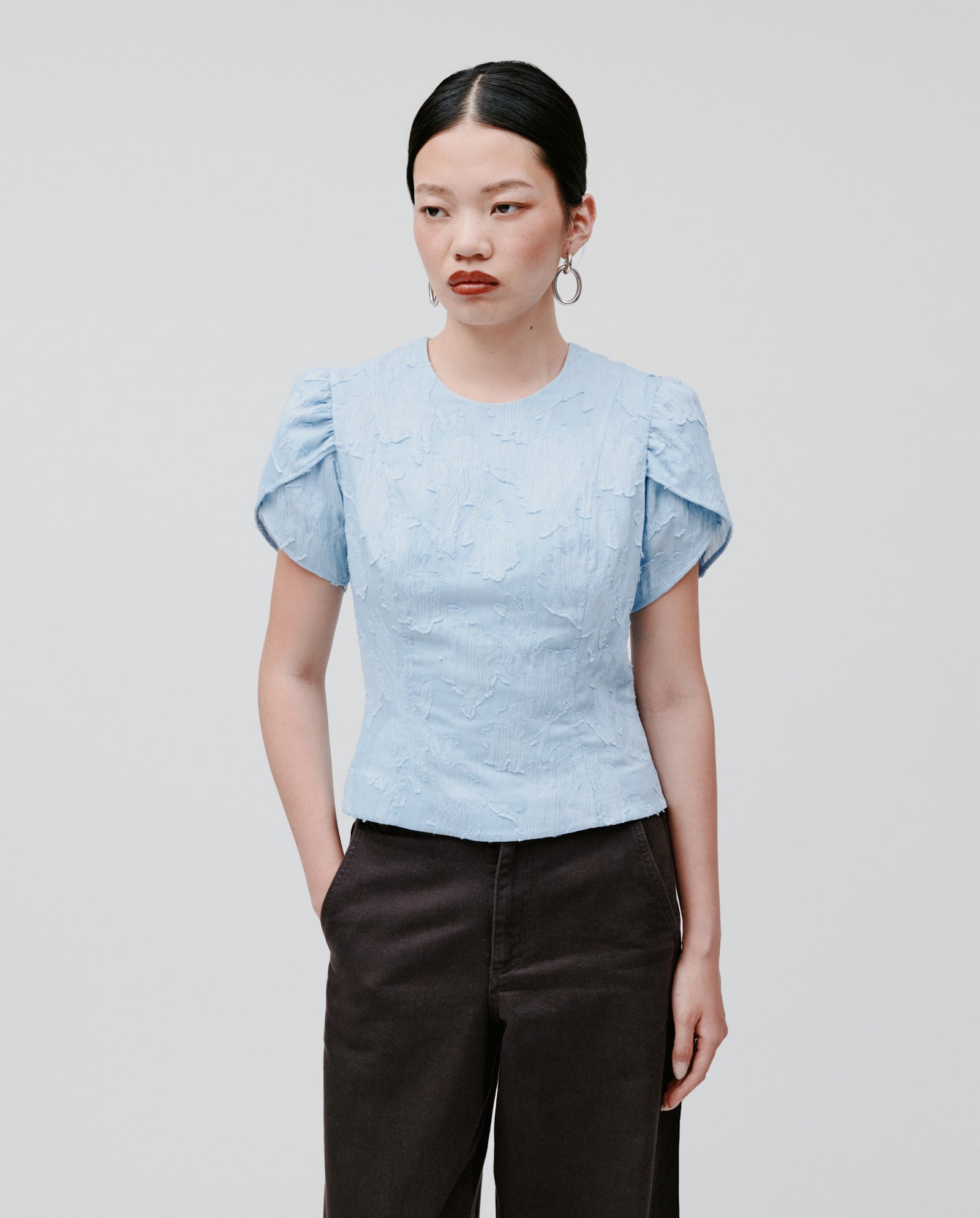 A woman wears the IVY OAK ELODIE Blouse, featuring a light blue fil coupé fabric, romantic silhouette, and short puff sleeves, paired with dark pants against a plain background.