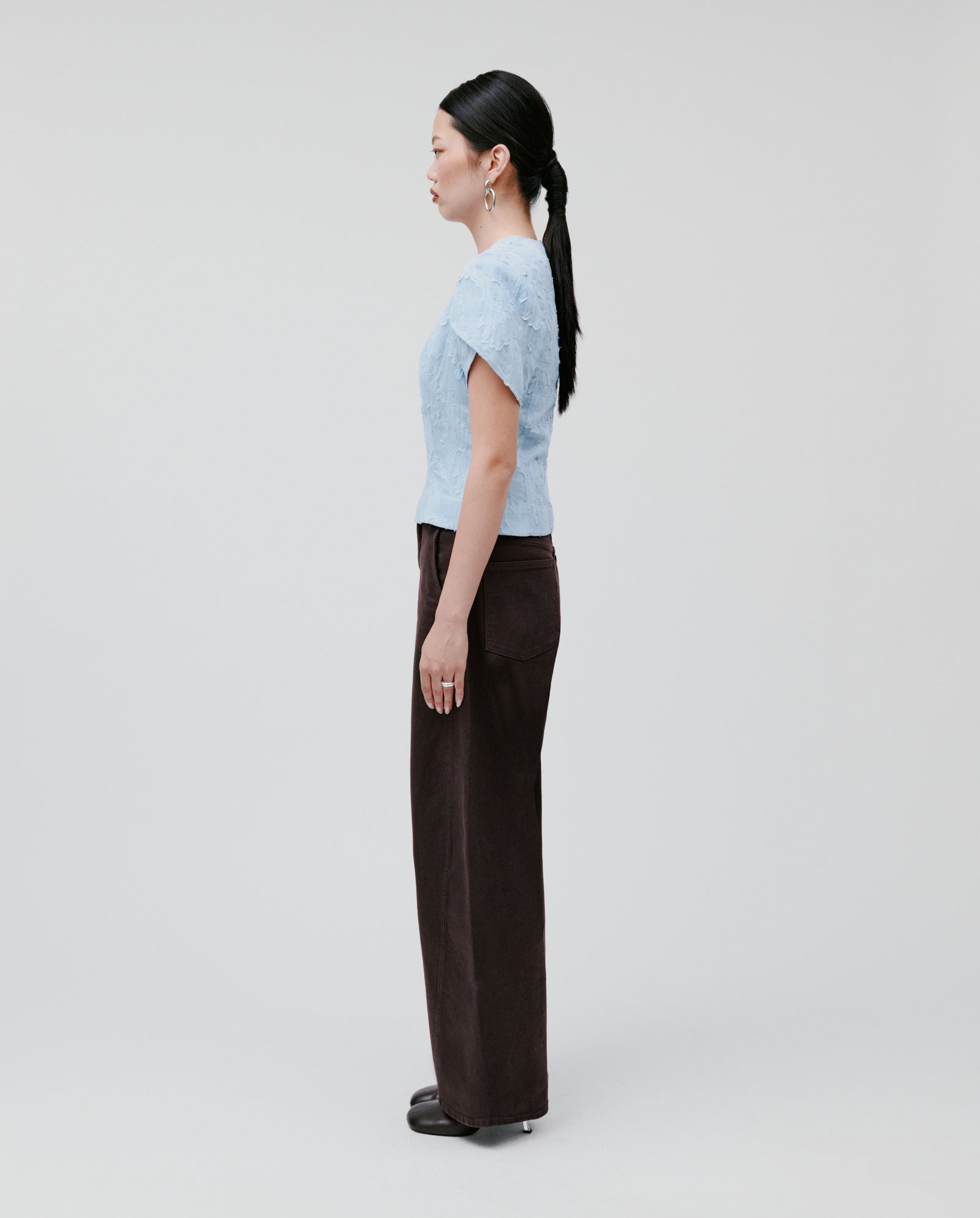 Woman wearing the ELODIE Blouse by IVY OAK, featuring fil coupé fabric and voluminous sleeves, paired with dark pants and standing in profile against a plain light background.