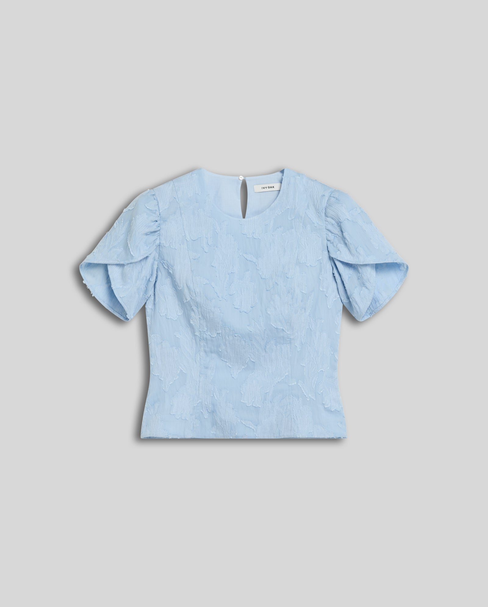 The ELODIE blouse by IVY OAK features light blue textured fil coupé fabric, romantic ruffled puff sleeves, and a flattering silhouette, showcased against a plain gray background.