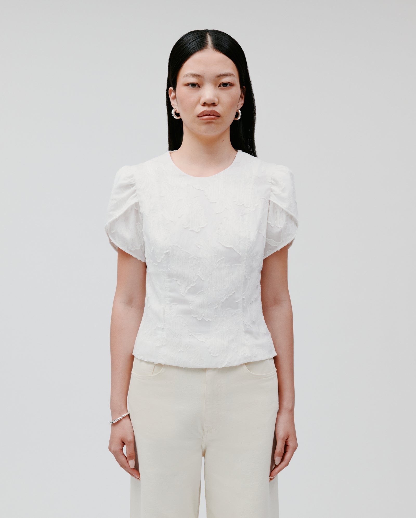 A woman with long black hair wears the IVY OAK ELODIE Blouse—white with fil coupé and voluminous sleeves—paired with cream pants, standing against a plain background.