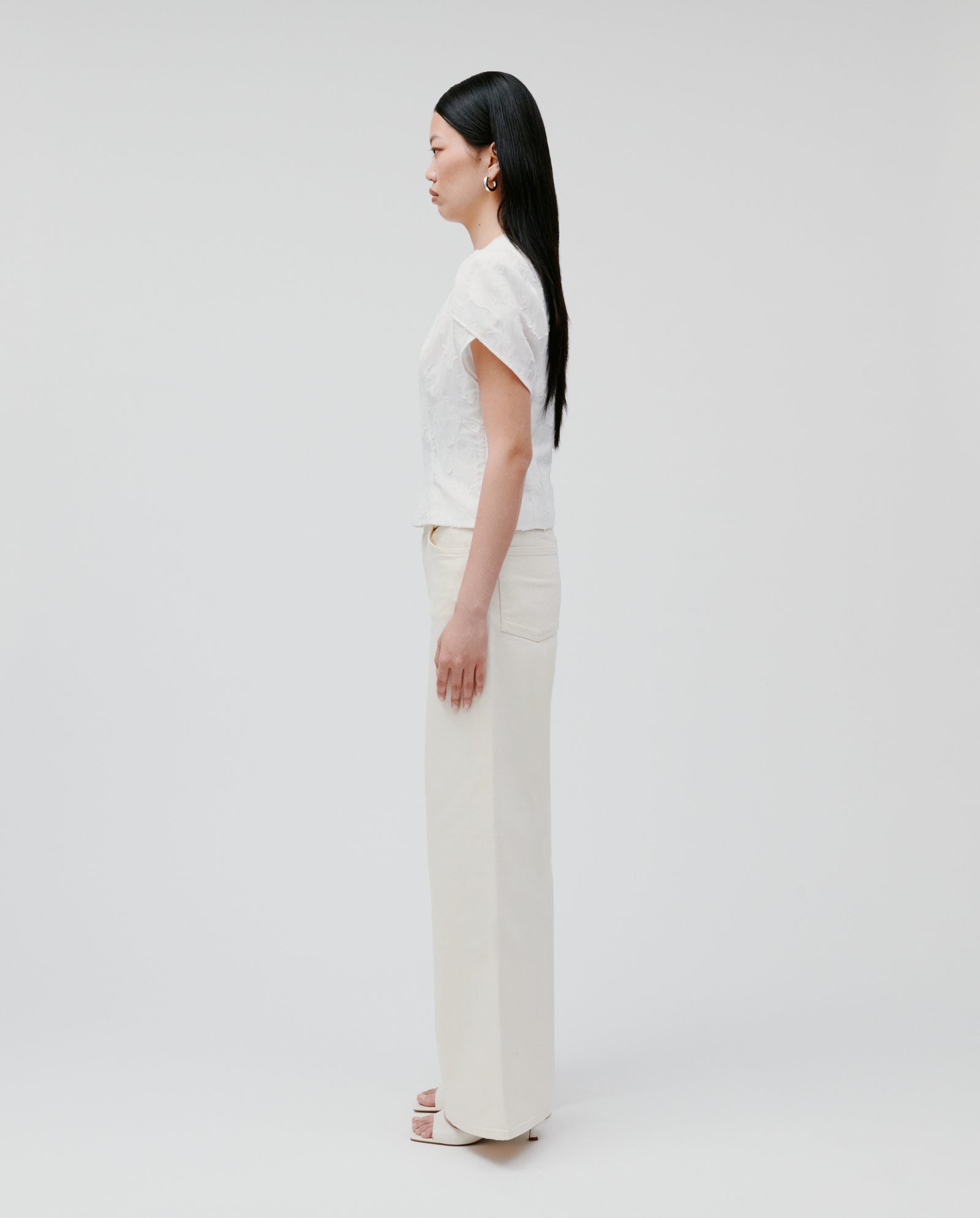 A woman stands in profile against a plain white background, wearing the IVY OAK ELODIE Blouse—a white short-sleeve top—paired with cream pants for a modern romantic silhouette.